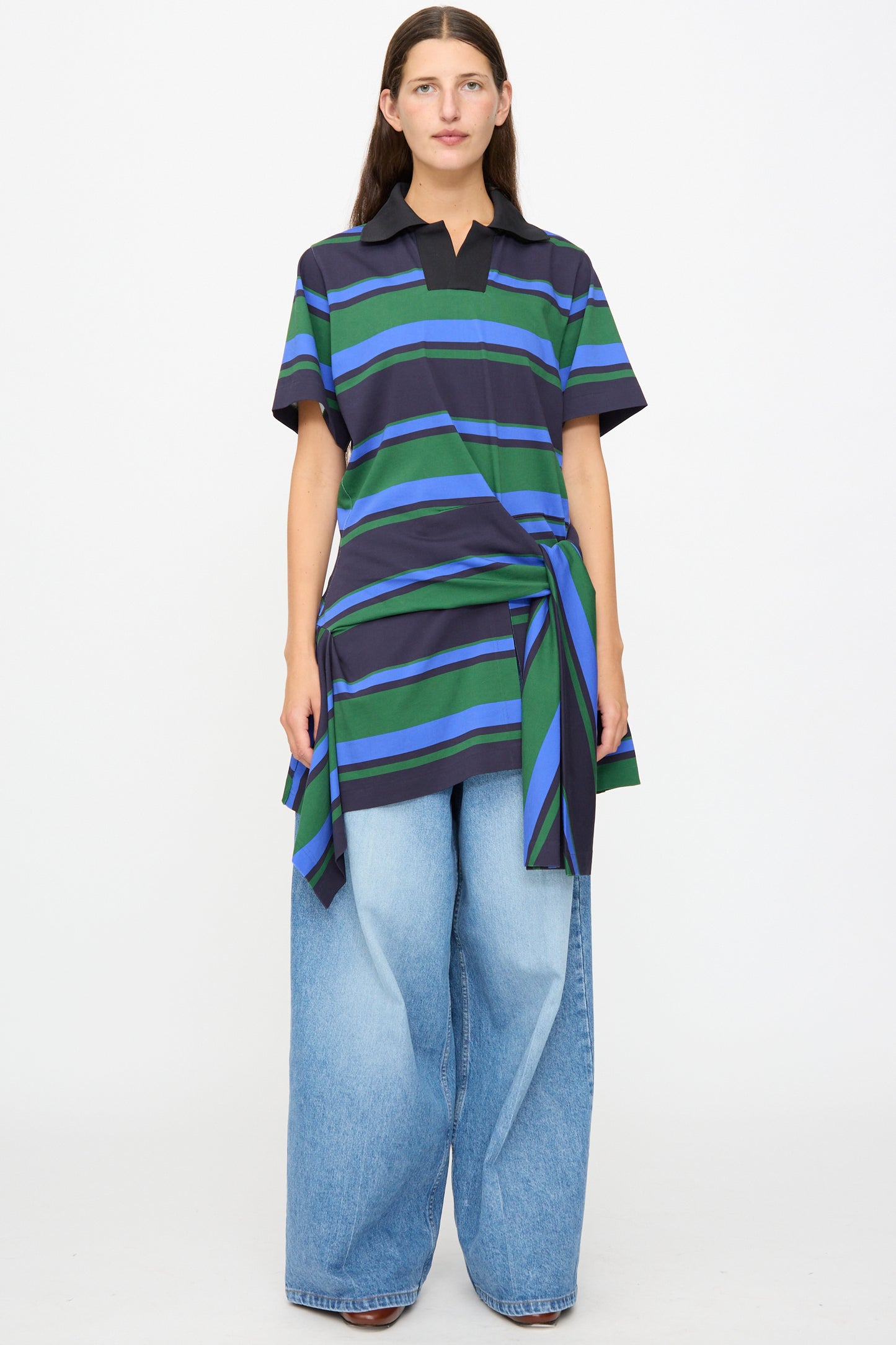 A person wearing the TOGA ARCHIVES Border Print Dress in Navy—a black, blue, and green striped cotton mini dress with an asymmetrical hem—paired with wide-leg light blue jeans, standing against a plain white background.