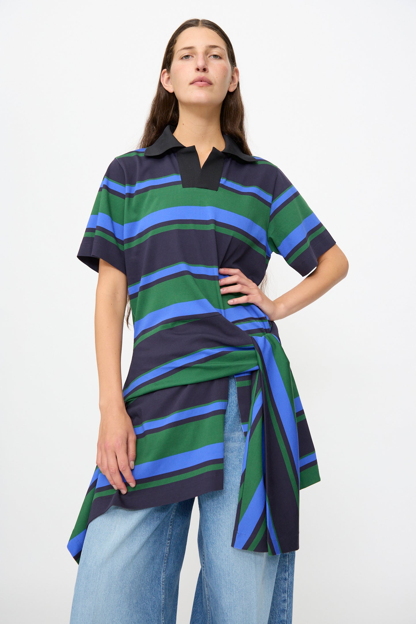 A person wearing the TOGA ARCHIVES Border Print Dress in Navy— a loose-fitting, short-sleeve tunic featuring green, blue, and black stripes— over wide-leg blue jeans, standing against a plain white background.