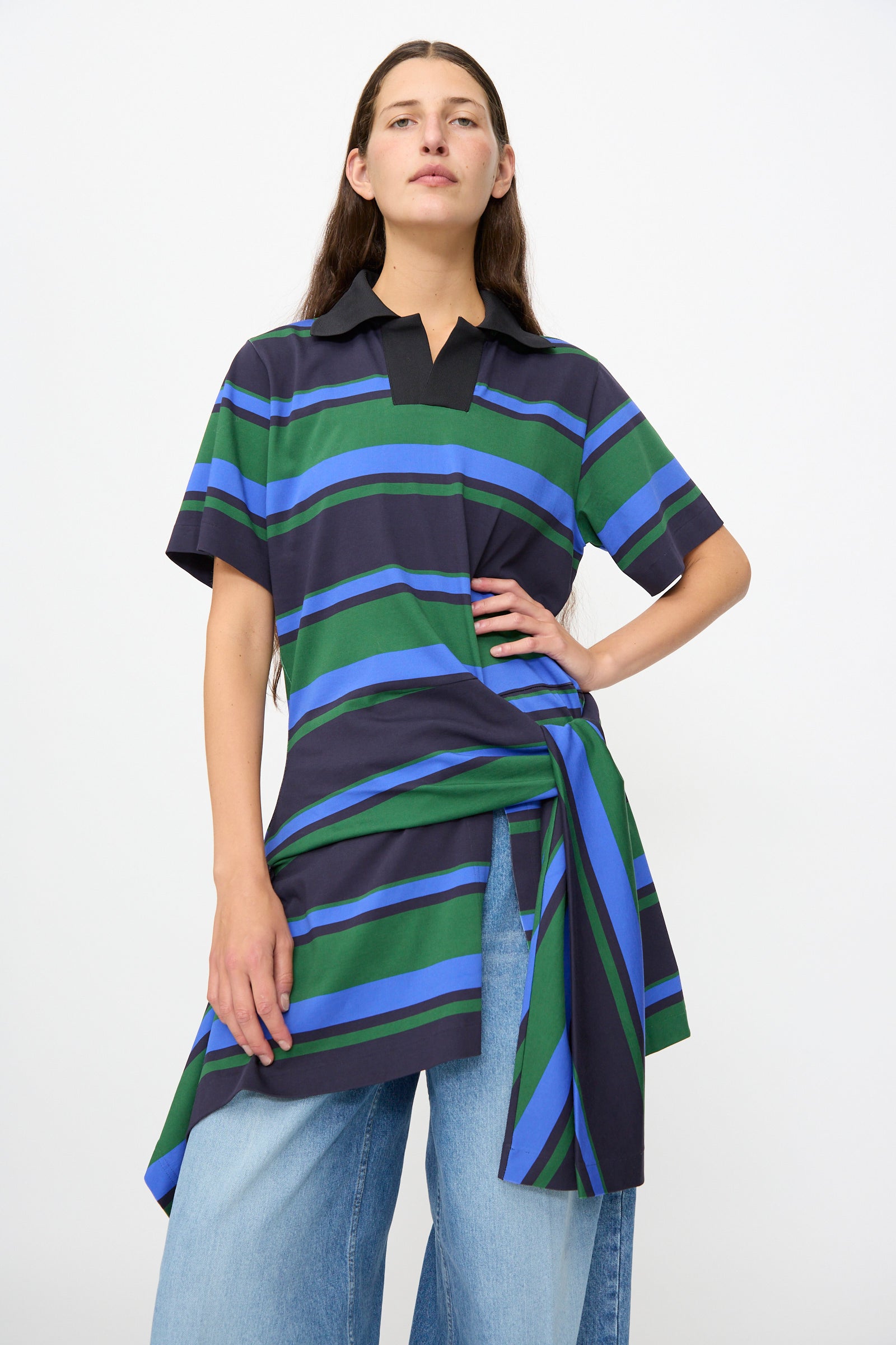 A person wearing the TOGA ARCHIVES Border Print Dress in Navy— a loose-fitting, short-sleeve tunic featuring green, blue, and black stripes— over wide-leg blue jeans, standing against a plain white background.
