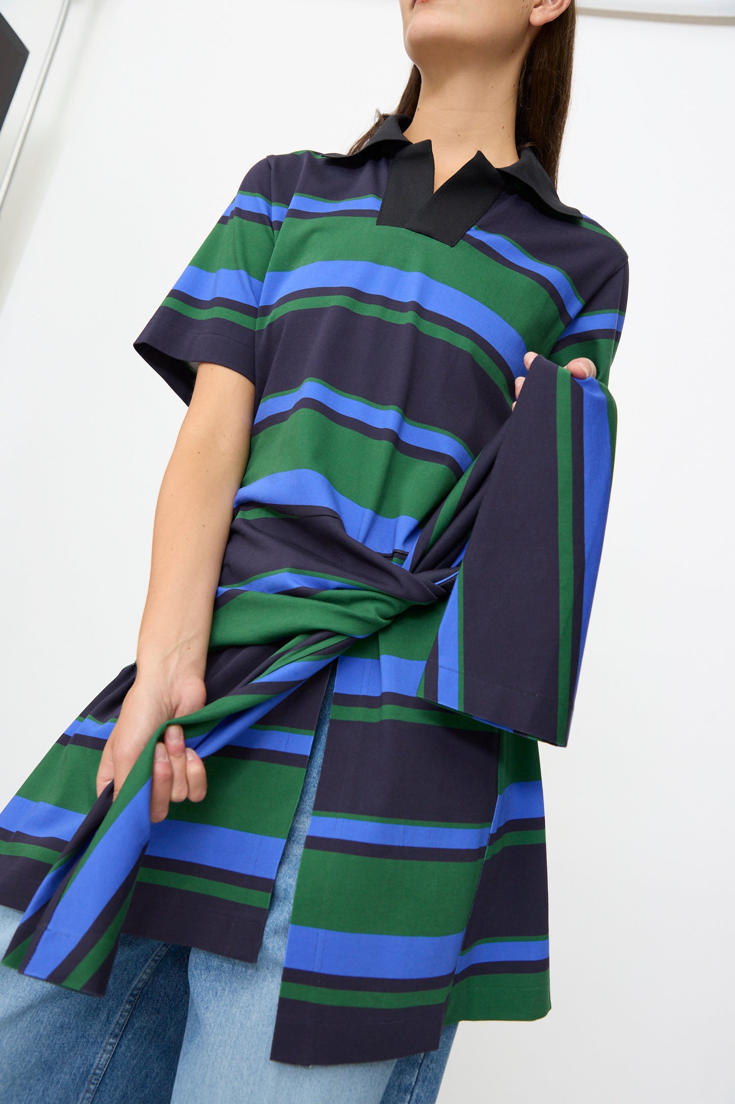 A person wearing the TOGA ARCHIVES Border Print Dress in Navy—a short-sleeve, striped cotton mini dress in shades of blue, green, and black—ties a loose fabric belt at the front for a flattering waist-defining look, styled with blue jeans.