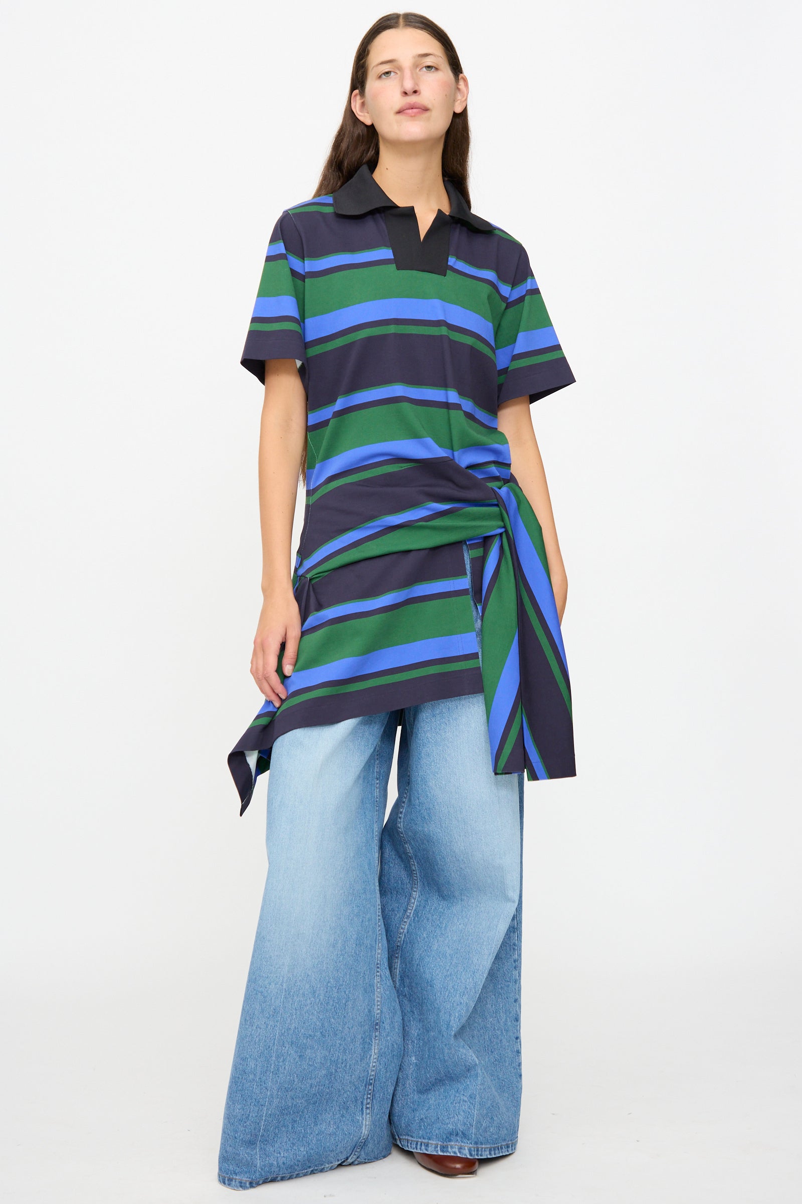 A person wearing the TOGA ARCHIVES Border Print Dress in Navy—a short-sleeve, striped cotton mini dress featuring an asymmetrical tie waist detail—styled over wide-leg light blue jeans, stands against a plain white background.