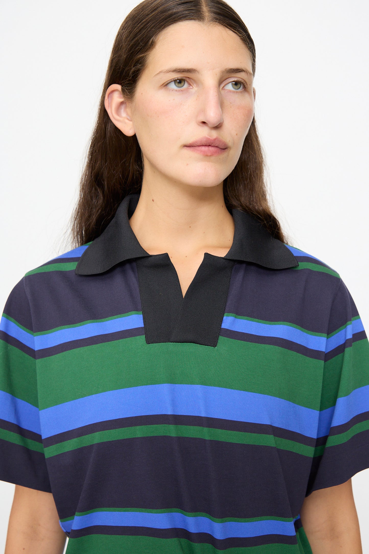 A woman with long brown hair wears the TOGA ARCHIVES Border Print Dress in Navy—a short-sleeve dress with a polo collar and blue, green, and black stripes—while looking slightly to the side against a plain white background.