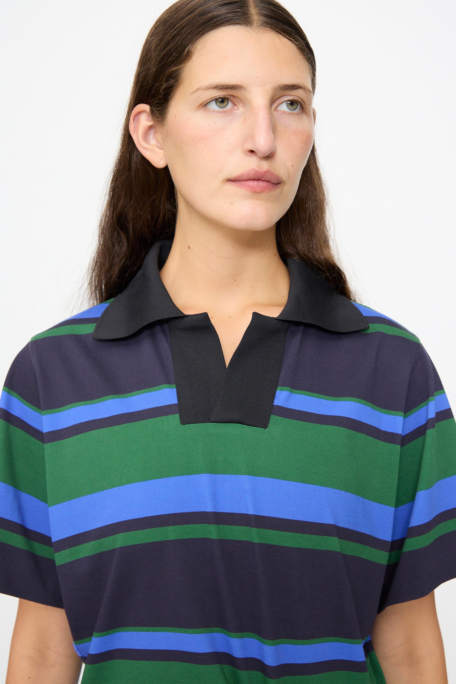 A woman with long brown hair wears the TOGA ARCHIVES Border Print Dress in Navy—a short-sleeve dress with a polo collar and blue, green, and black stripes—while looking slightly to the side against a plain white background.