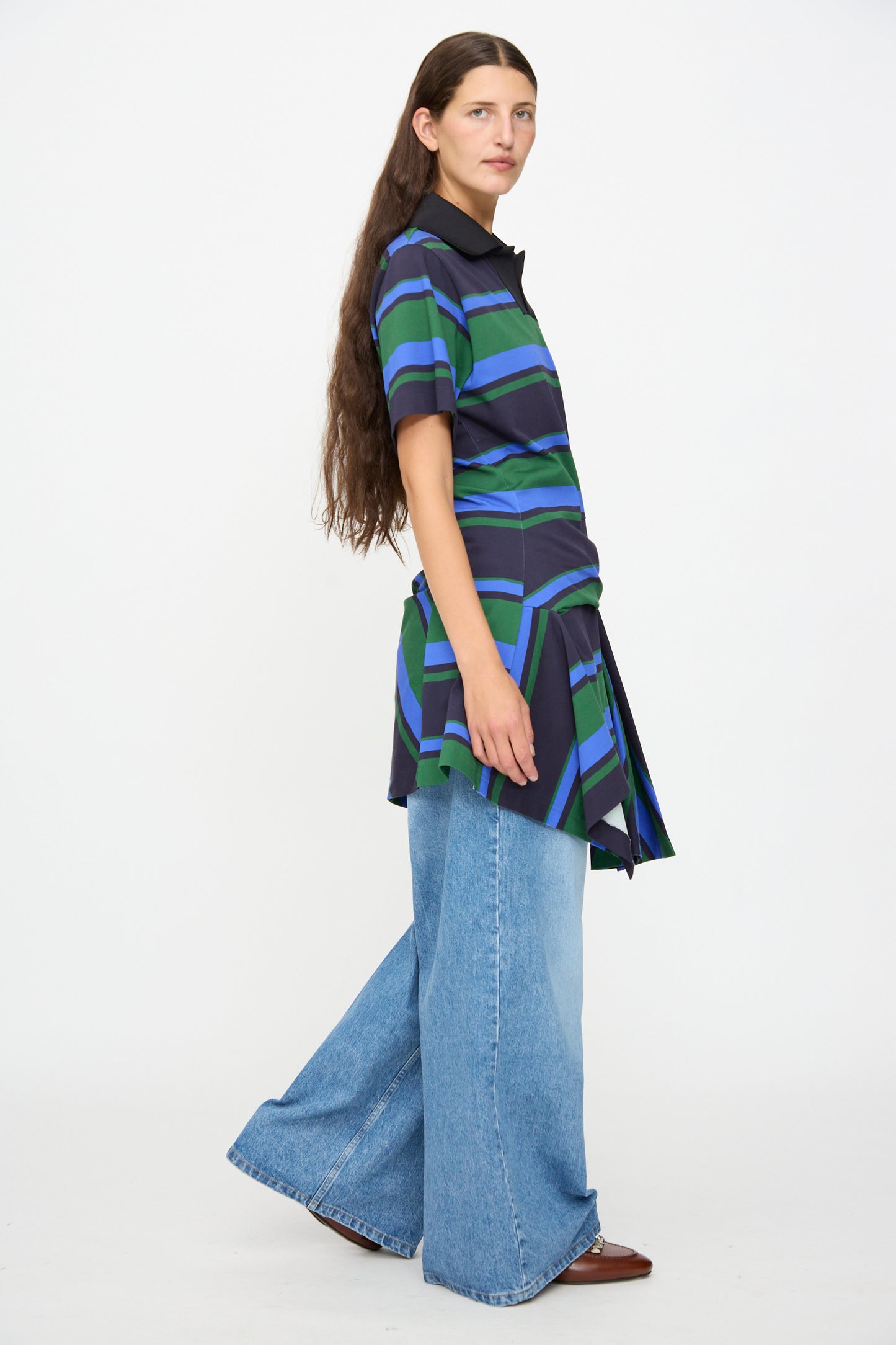 A woman with long brown hair wears the Border Print Dress in Navy by TOGA ARCHIVES, styled as a short-sleeved tunic top over wide-leg blue jeans and brown shoes, standing against a plain white background.