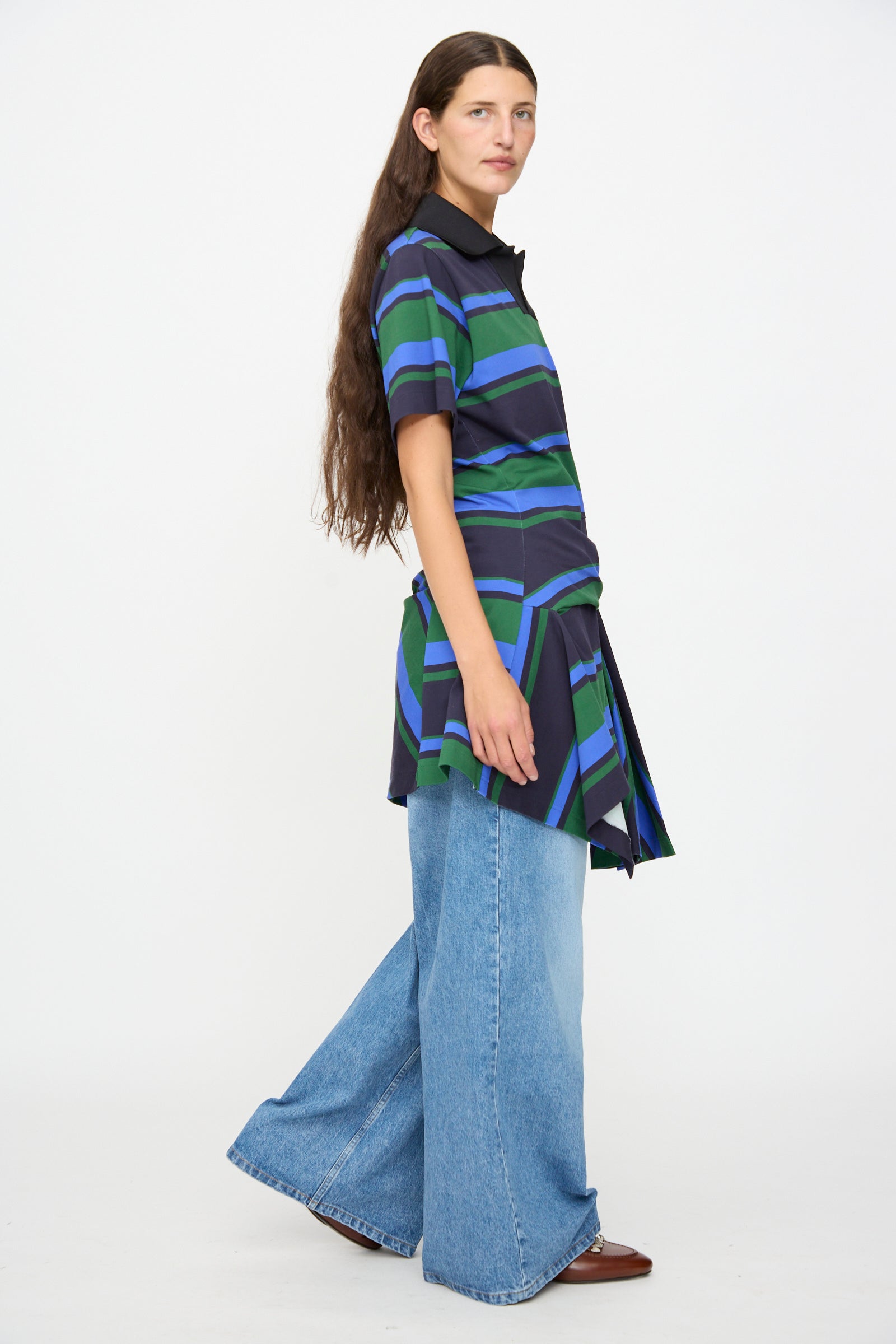 A woman with long brown hair wears the Border Print Dress in Navy by TOGA ARCHIVES, styled as a short-sleeved tunic top over wide-leg blue jeans and brown shoes, standing against a plain white background.