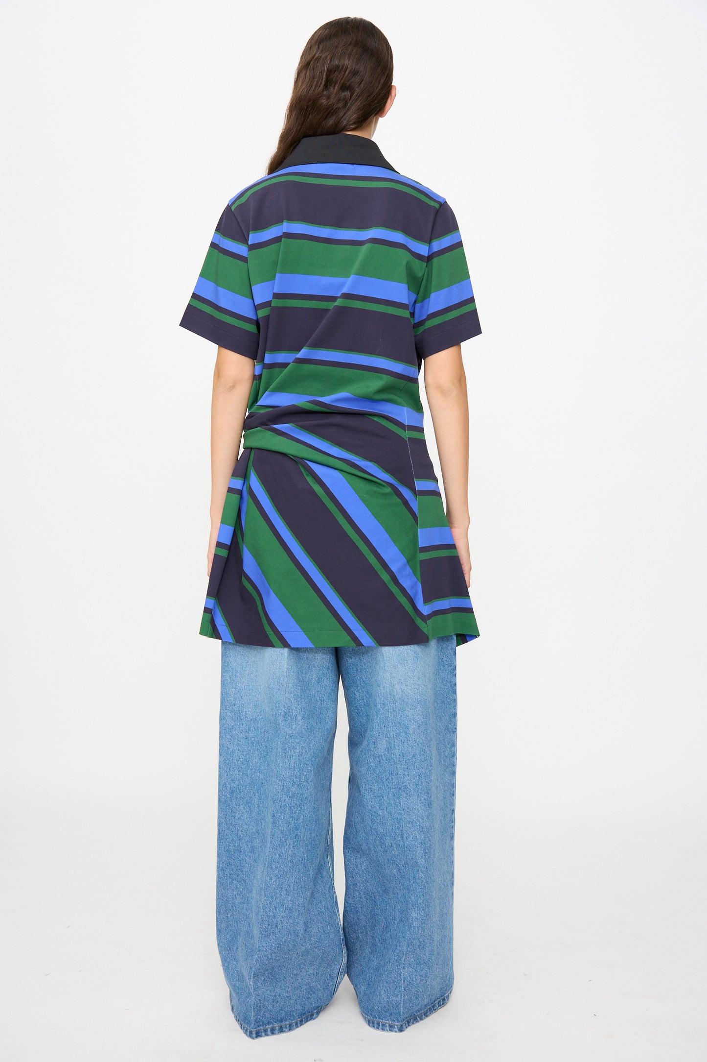 A person stands with their back to the camera, wearing the TOGA ARCHIVES Border Print Dress in Navy— a short-sleeve cotton mini dress featuring navy, green, and blue stripes — styled over wide-leg light blue jeans against a plain white background.
