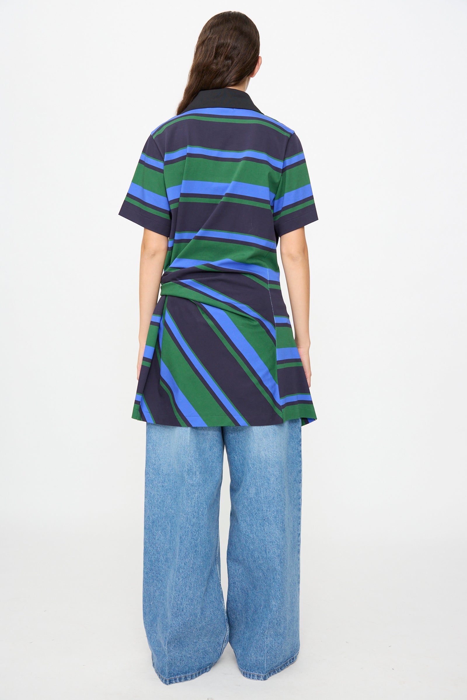 A person stands with their back to the camera, wearing the TOGA ARCHIVES Border Print Dress in Navy— a short-sleeve cotton mini dress featuring navy, green, and blue stripes — styled over wide-leg light blue jeans against a plain white background.