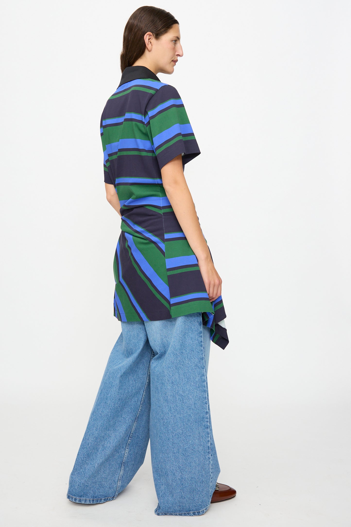 A person stands sideways against a plain white background wearing the TOGA ARCHIVES Border Print Dress in Navy—a short-sleeve, blue and green striped cotton mini dress—layered over wide-leg blue jeans and brown shoes.