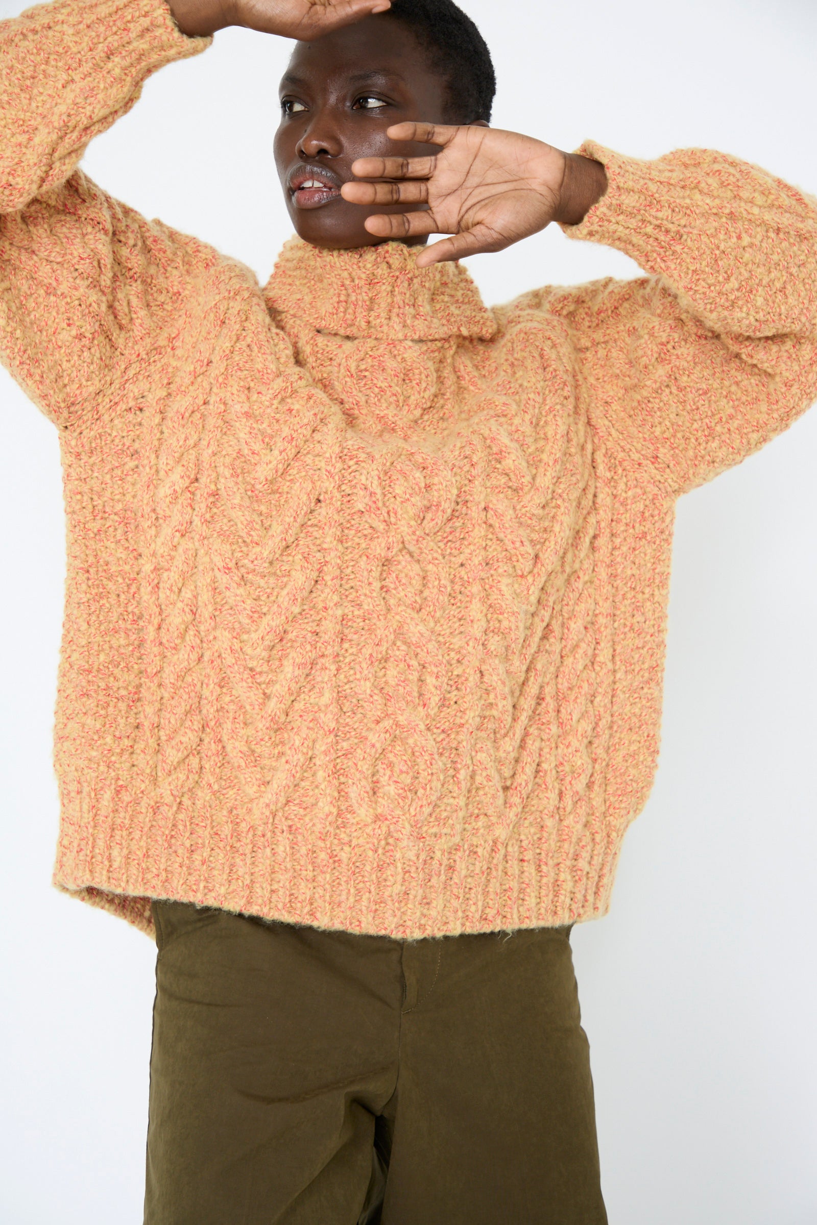 A person wearing a textured, yellow Cable Knit Pullover from TOGA ARCHIVES and khaki pants, with arms raised against a plain background.