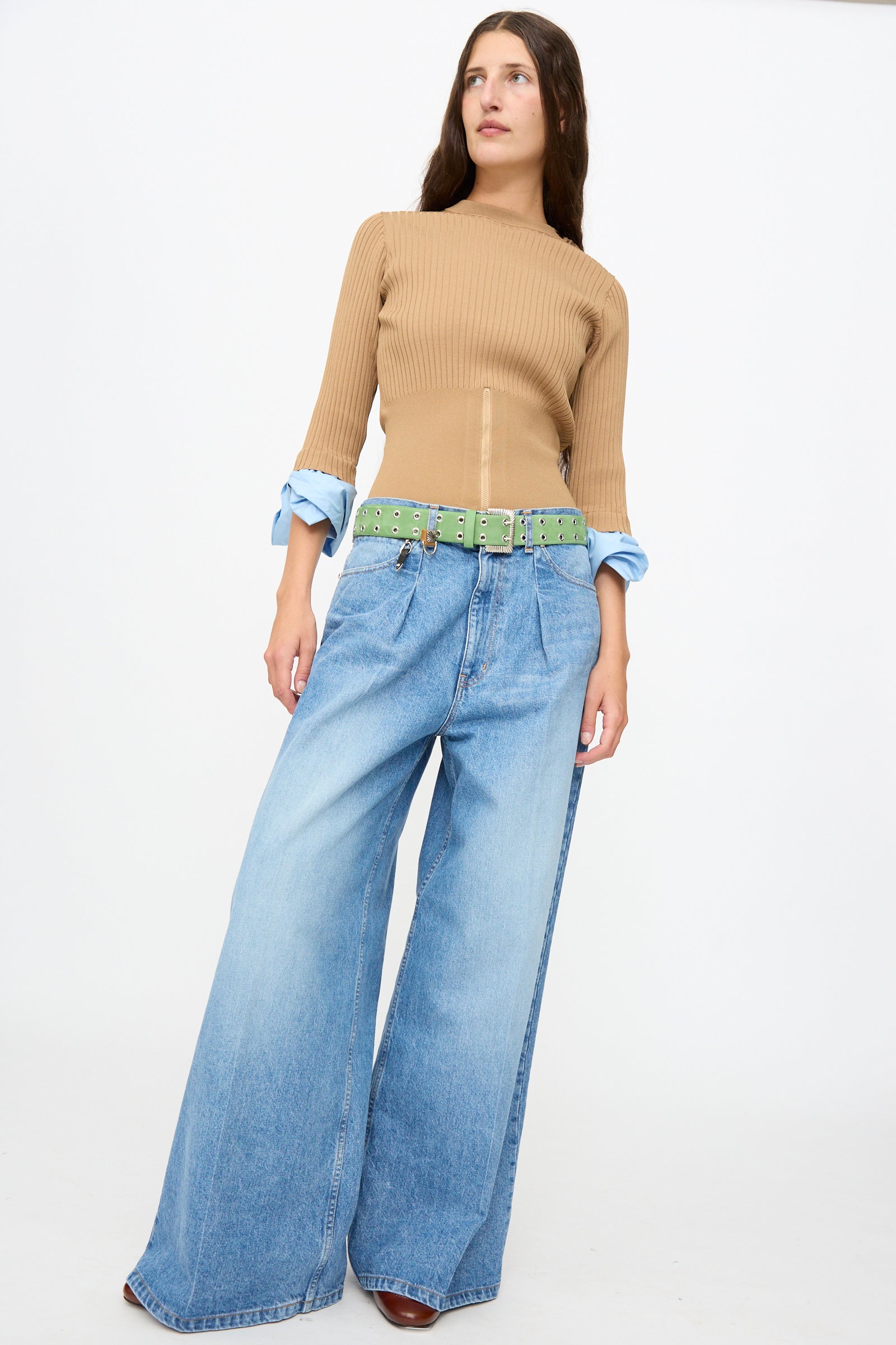 A person stands against a white background wearing a tan ribbed sweater, light blue wide-leg jeans, the Eyelet Metal Belt in Light Green by TOGA ARCHIVES, and brown shoes.