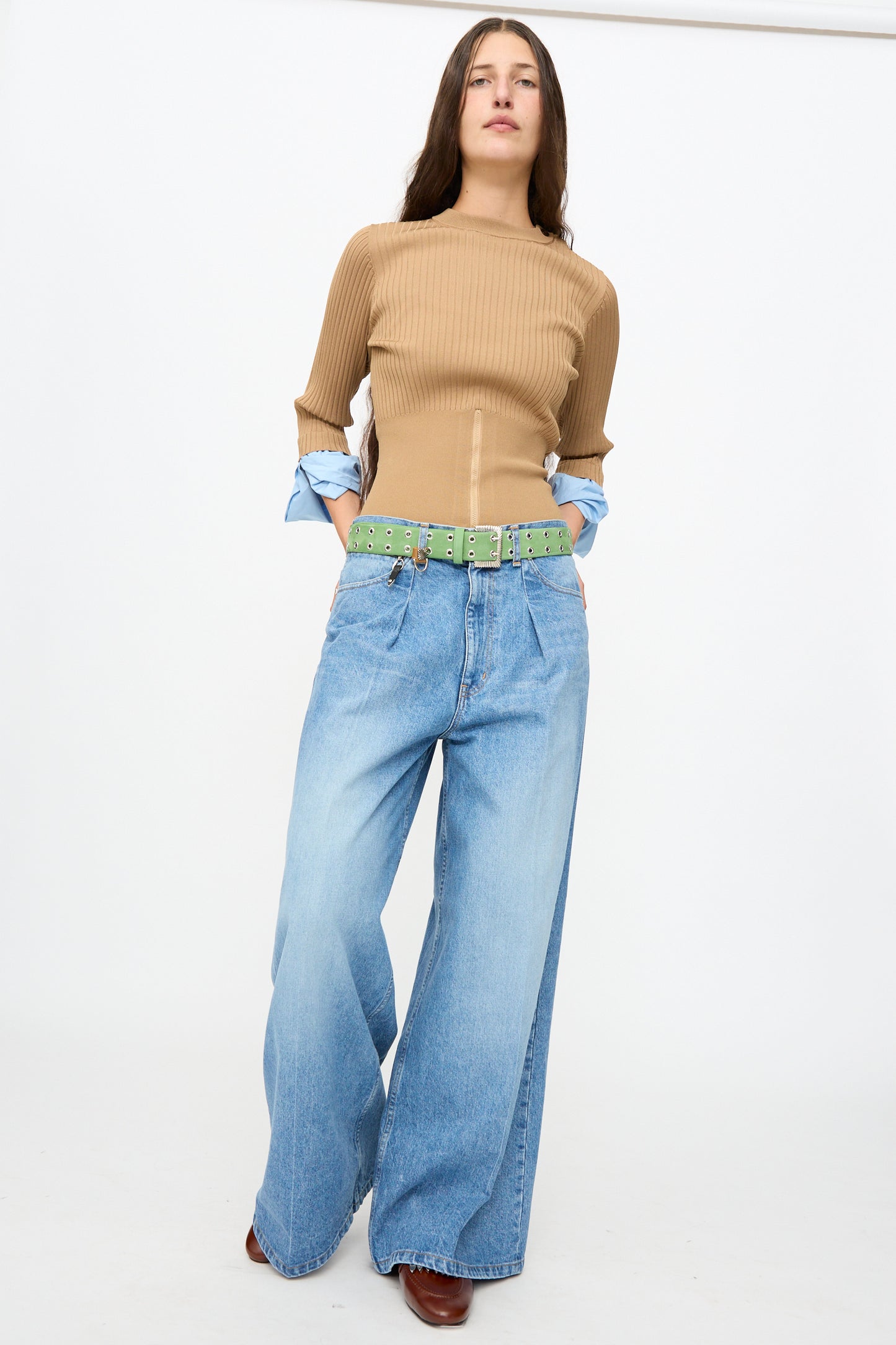Person standing against a white background wearing a tan ribbed top, light blue wide-leg jeans, and brown shoes, styled with the TOGA ARCHIVES Eyelet Metal Belt in Light Green made in Japan, with hands in pockets.
