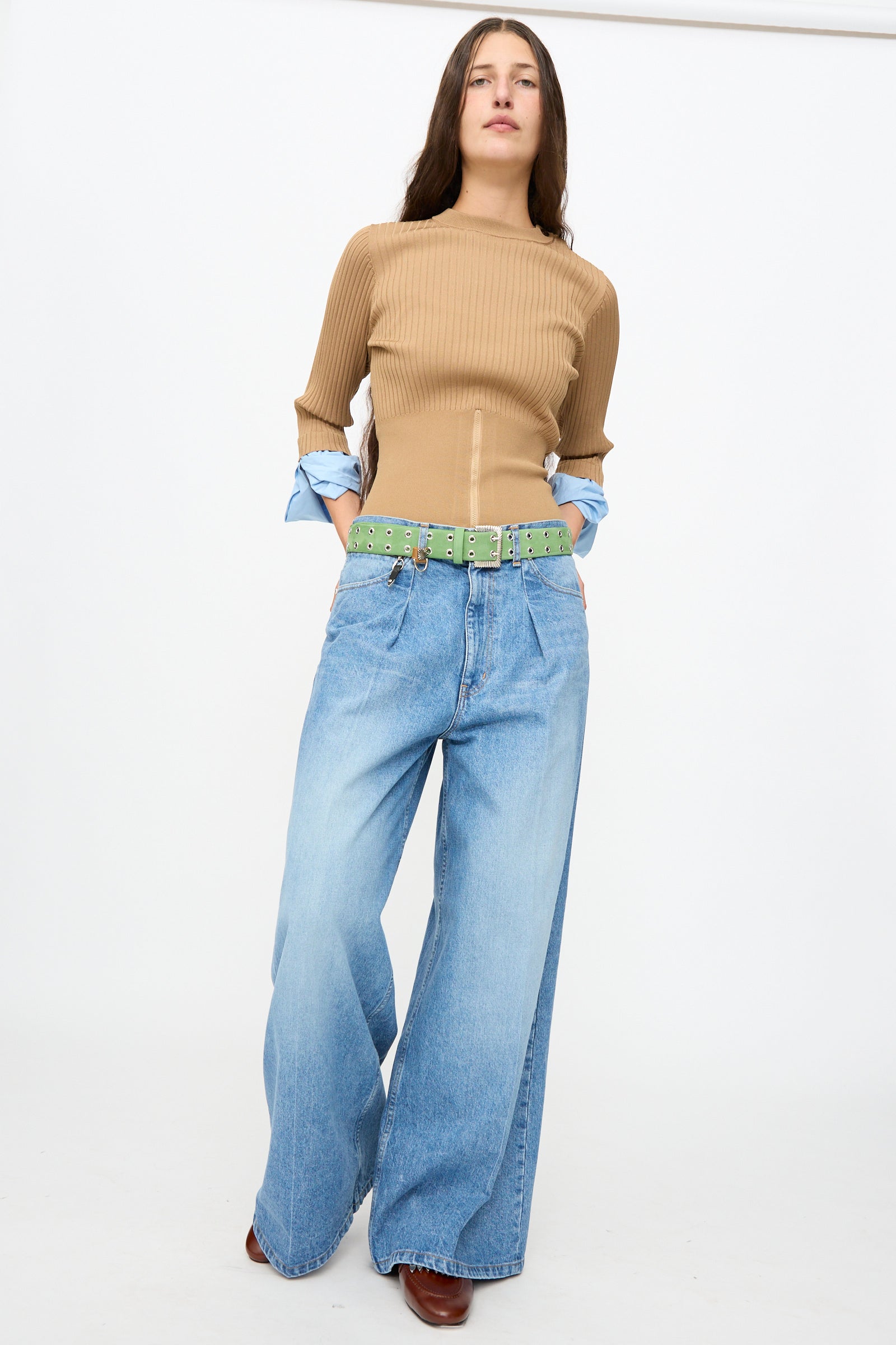 Person standing against a white background wearing a tan ribbed top, light blue wide-leg jeans, and brown shoes, styled with the TOGA ARCHIVES Eyelet Metal Belt in Light Green made in Japan, with hands in pockets.