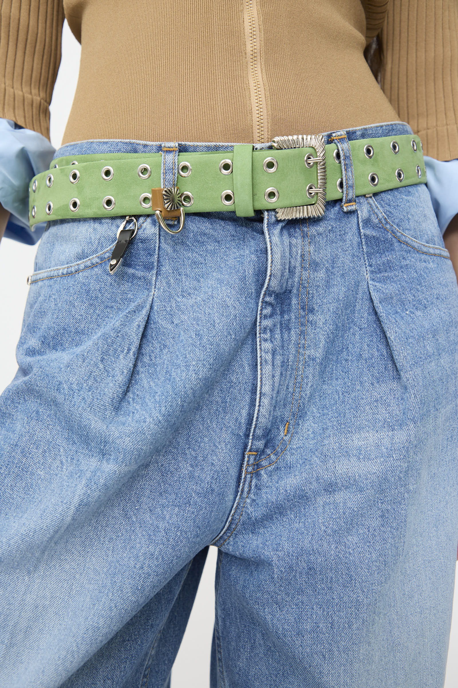 Wearing light blue jeans paired with the TOGA ARCHIVES Eyelet Metal Belt in Light Green, this person also has on a tan ribbed top and partially visible blue shirt sleeves; the belt features distinctive metal hardware including a small lock and key.