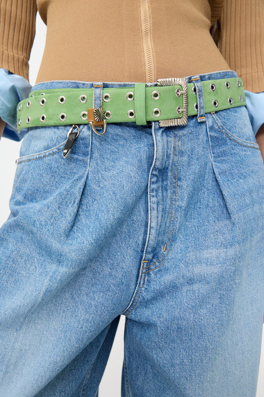 Wearing light blue jeans paired with the TOGA ARCHIVES Eyelet Metal Belt in Light Green, this person also has on a tan ribbed top and partially visible blue shirt sleeves; the belt features distinctive metal hardware including a small lock and key.