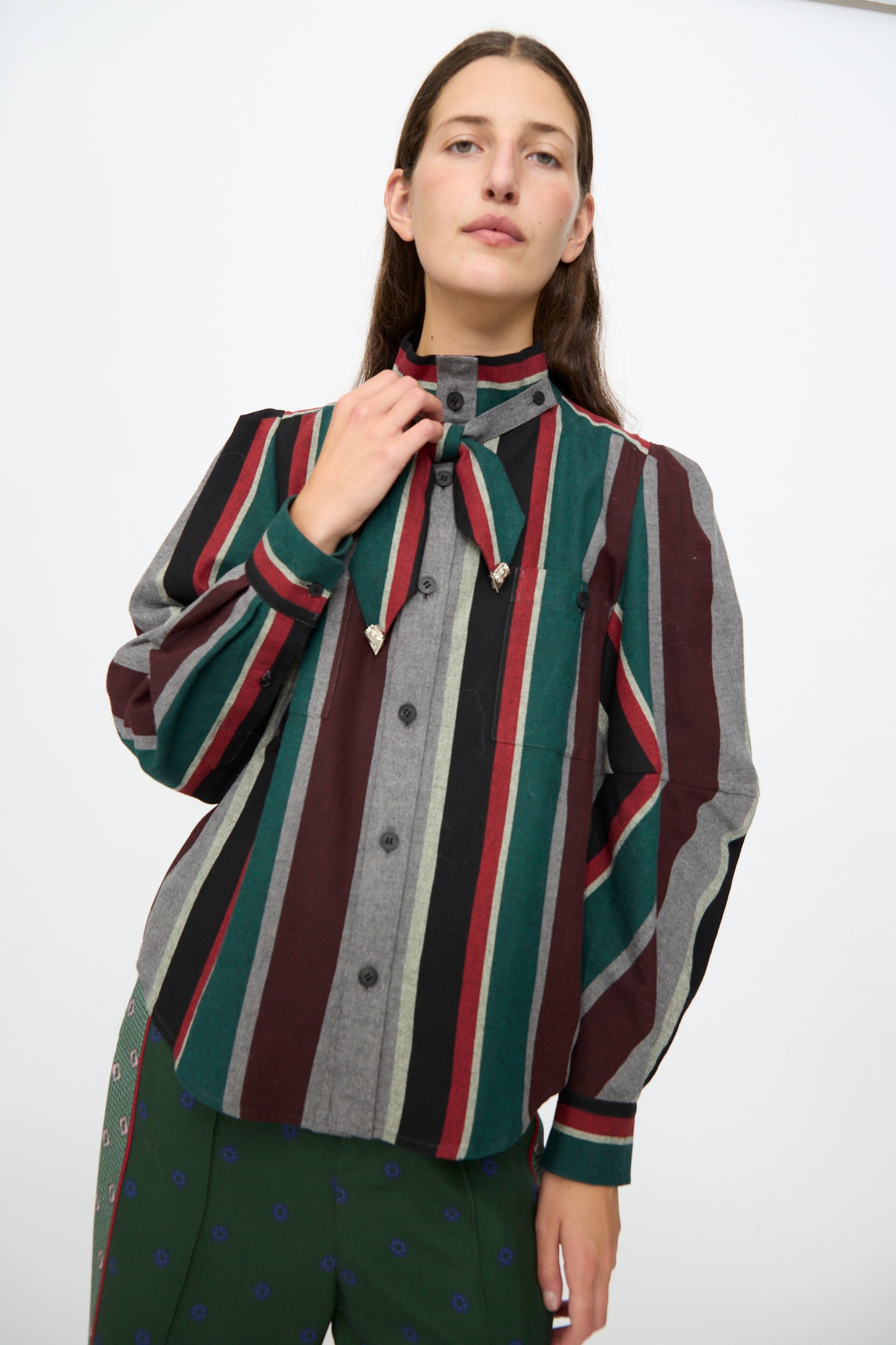 A person is wearing the TOGA ARCHIVES Flannel Striped Shirt in Green—a long sleeve, button-up cotton shirt featuring vertical stripes in red, green, gray, and black with a distinctive necktie detail—standing against a plain white background. This stylish piece is expertly made in Japan.