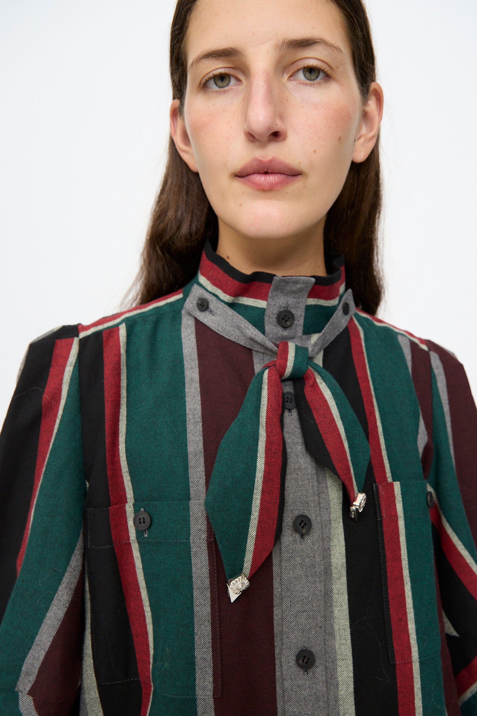 A person wearing the TOGA ARCHIVES Flannel Striped Shirt in Green, a long-sleeve cotton shirt with vertical green, red, and gray stripes and a coordinating bow at the collar. Made in Japan. The background is plain white.