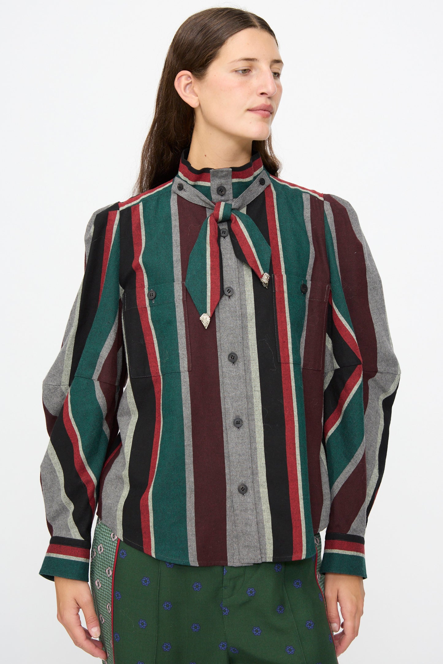 A person wearing the TOGA ARCHIVES Flannel Striped Shirt in Green— a long-sleeve, multi-colored vertical striped cotton shirt made in Japan with a necktie detail—paired with a subtly patterned green skirt and standing against a plain background.