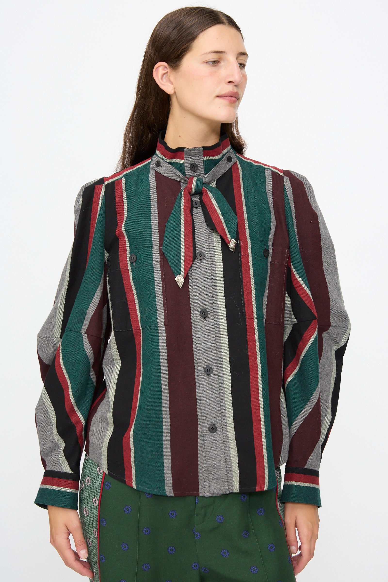 A person wearing the TOGA ARCHIVES Flannel Striped Shirt in Green— a long-sleeve, multi-colored vertical striped cotton shirt made in Japan with a necktie detail—paired with a subtly patterned green skirt and standing against a plain background.