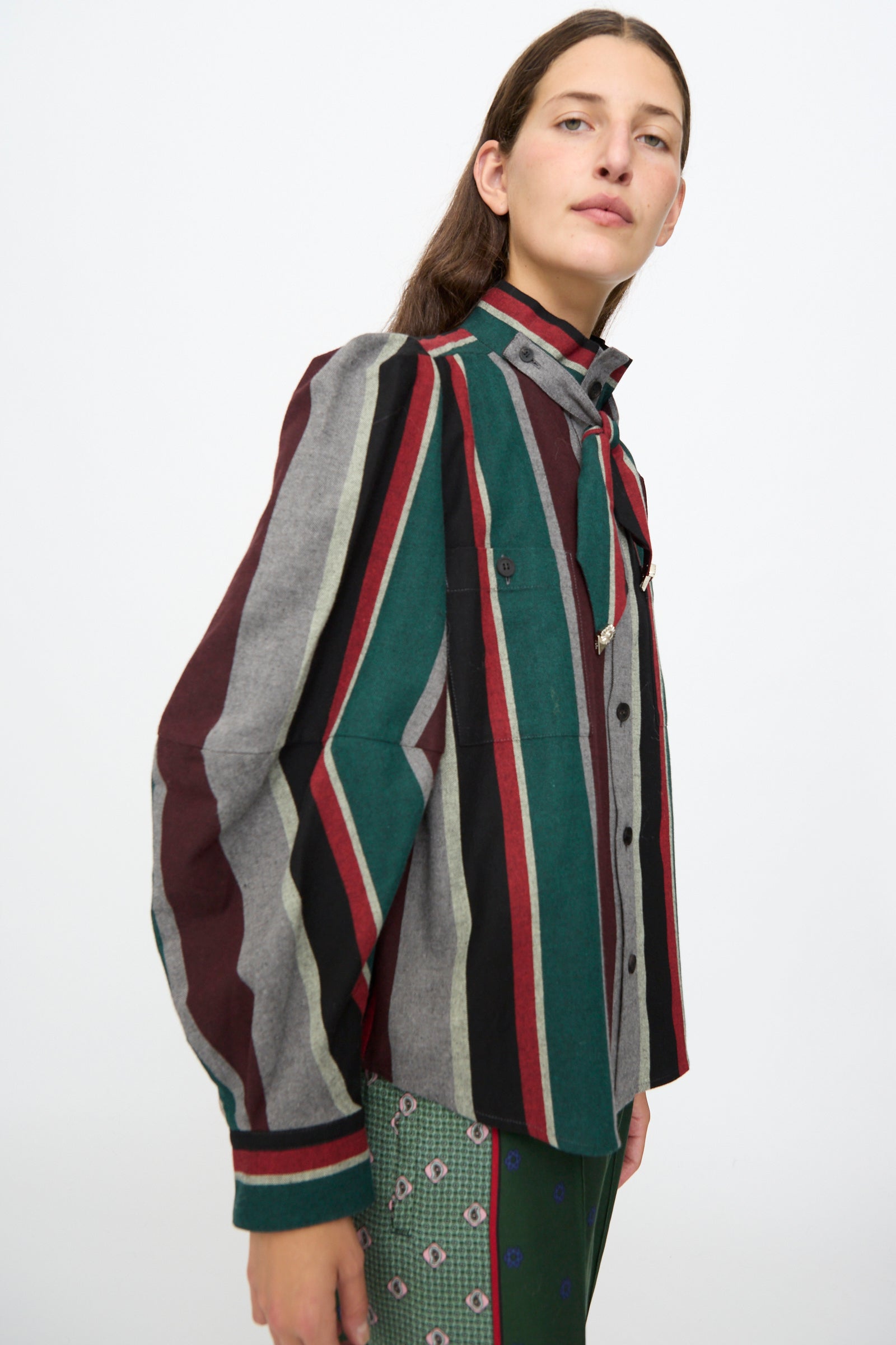 A person stands against a plain background, wearing the TOGA ARCHIVES Flannel Striped Shirt in Green, featuring vertical red, green, black, and gray stripes, paired with patterned green pants.