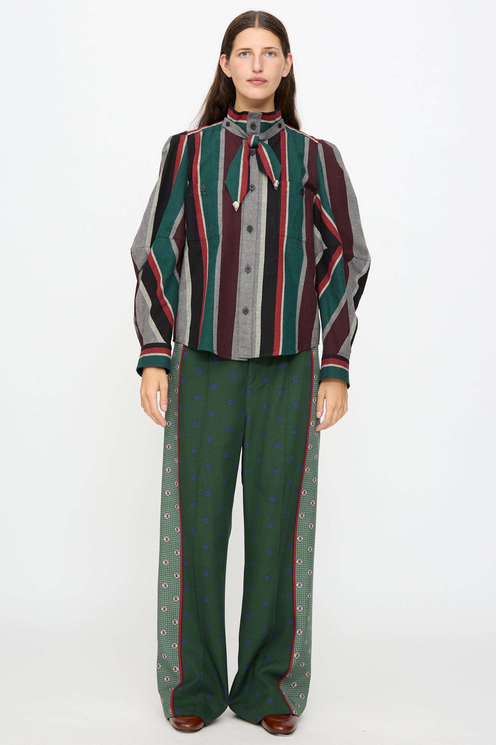 A person stands facing forward against a plain white background, wearing the TOGA ARCHIVES Flannel Striped Shirt in Green, a made-in-Japan cotton long sleeve top featuring a bow collar, paired with green patterned wide-leg pants.