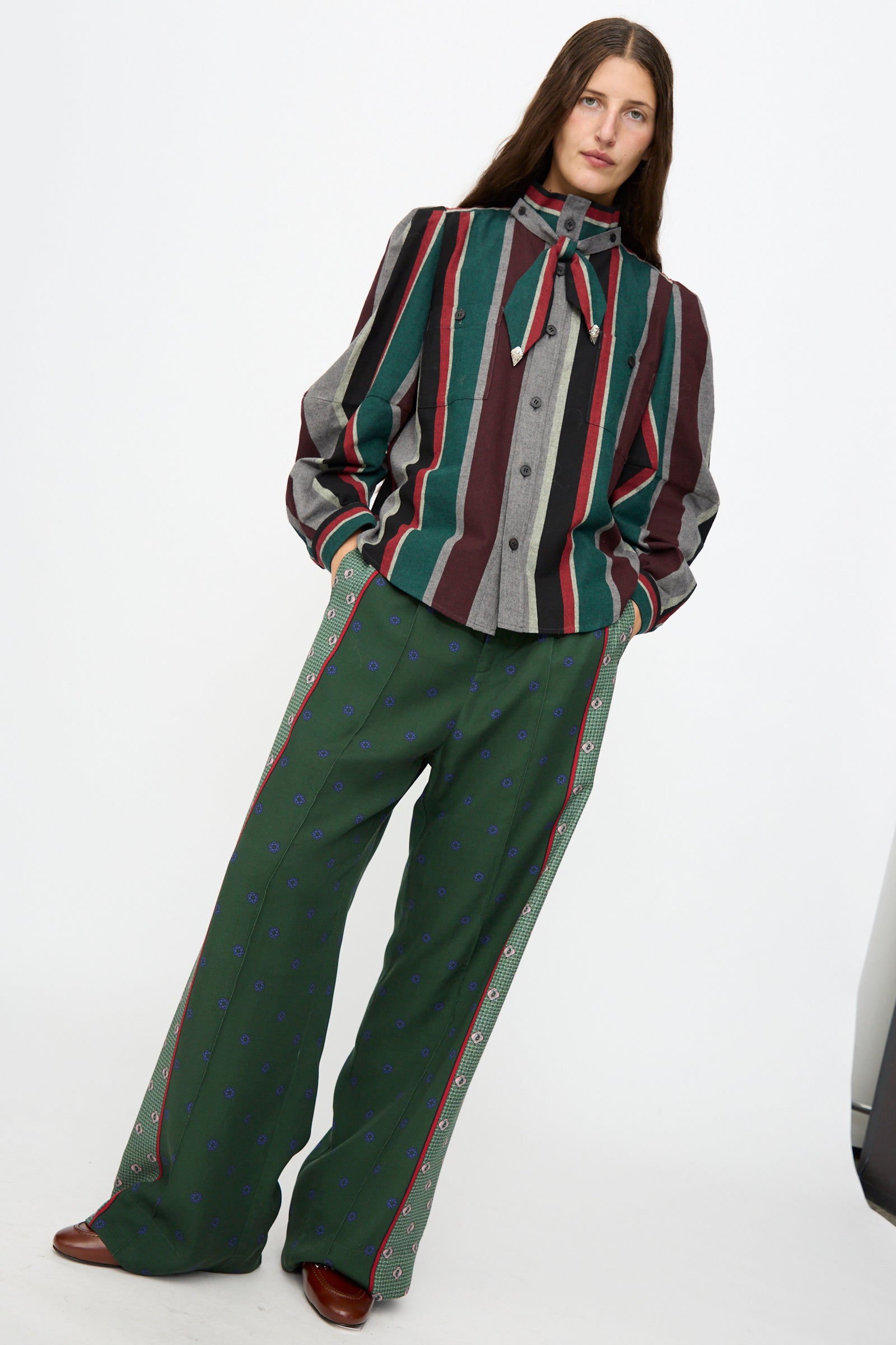 A person wearing the TOGA ARCHIVES Flannel Striped Shirt in Green, made in Japan, with long sleeves and a necktie, paired with wide-leg green pants adorned with small blue patterns, stands against a plain white background.