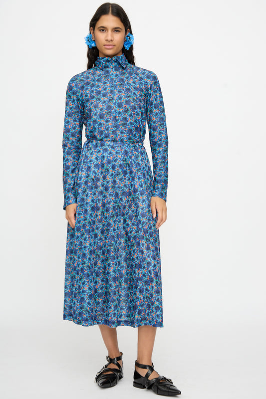 A woman stands against a white background, wearing the TOGA ARCHIVES Flared Jersey Dress in Mix—a long-sleeved midi dress with an adjustable waist—paired with black shoes and blue flower hair accessories.
