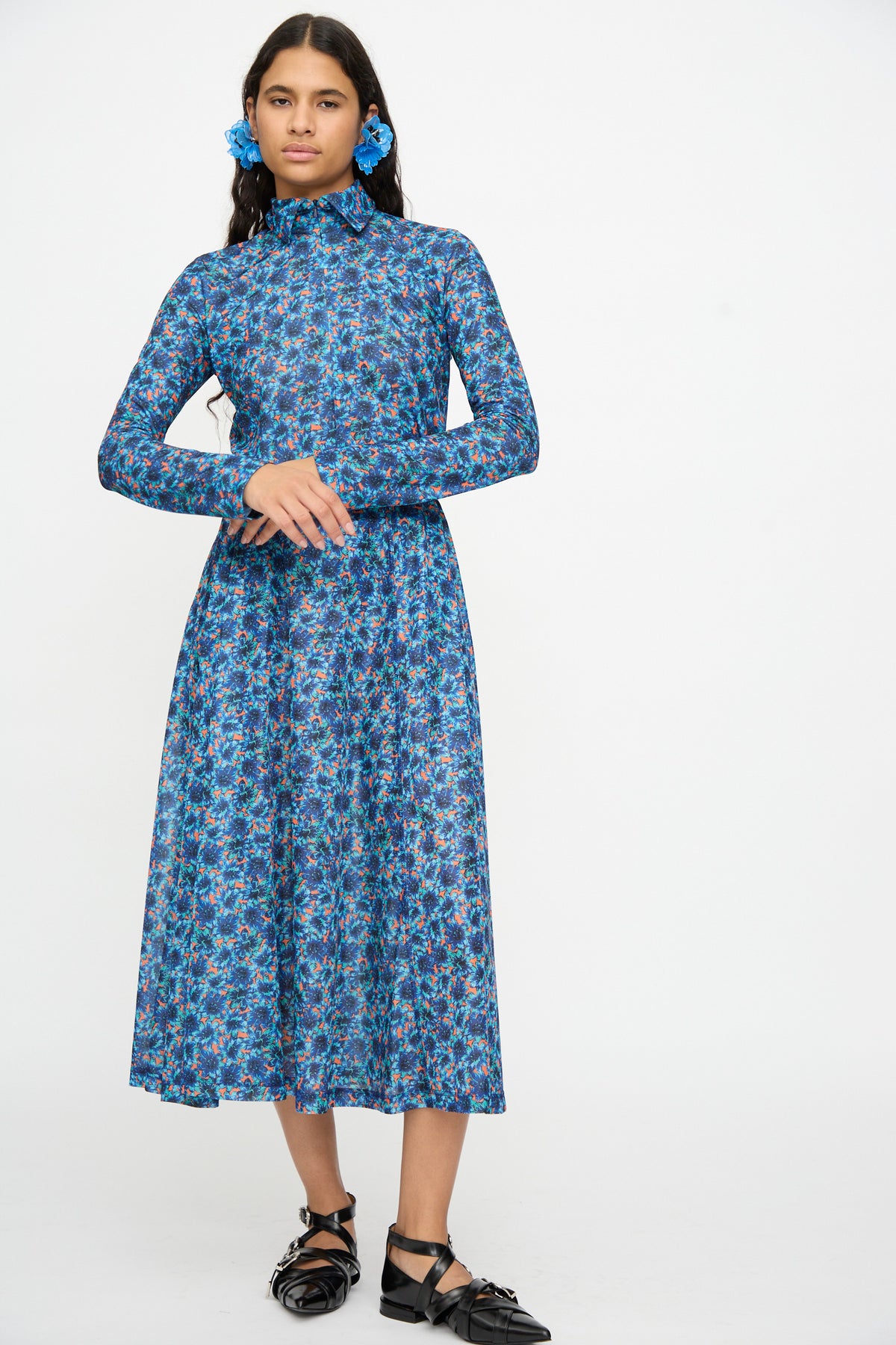 A person wearing the TOGA ARCHIVES Flared Jersey Dress in Mix, featuring long sleeves, a midi length, a high neck, and a floral print, with black sandals, stands against a plain white background.