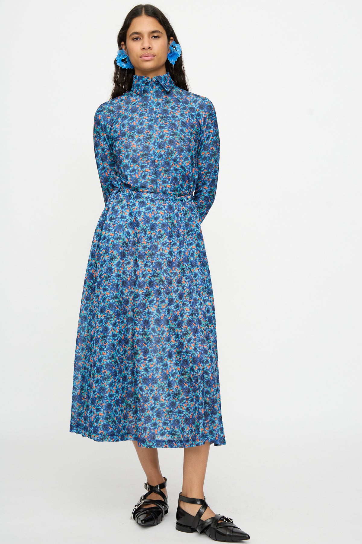 A woman stands against a white background wearing the TOGA ARCHIVES Flared Jersey Dress in Mix, a long-sleeved midi dress with a high neck and adjustable waist for comfort and style. She pairs the look with black flat shoes and blue floral hair accessories, her hands elegantly placed behind her back—an effortlessly elegant ensemble.