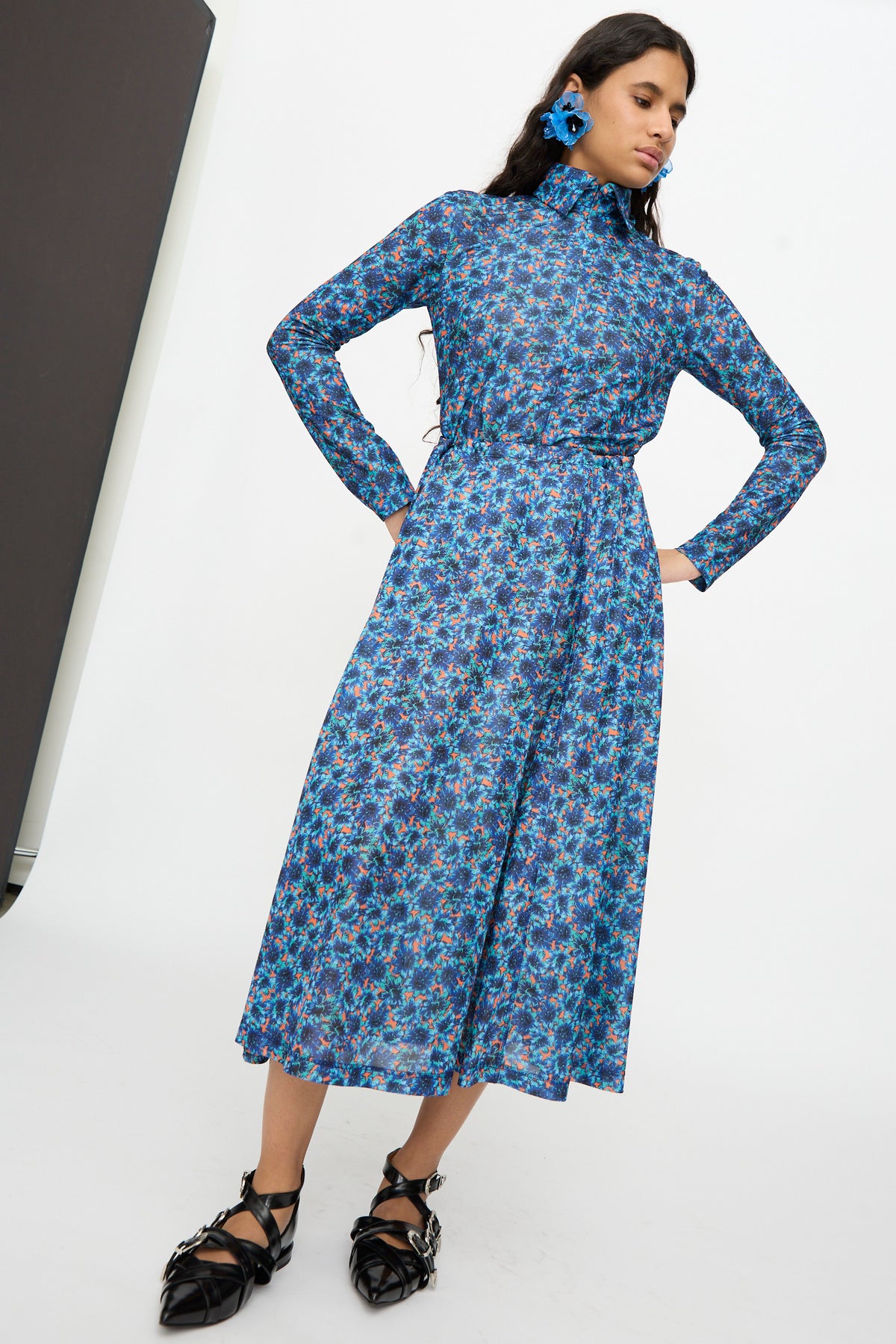 A woman wearing the TOGA ARCHIVES Flared Jersey Dress in Mix, styled with blue earrings and black shoes, stands against a white background with a black panel on the left.