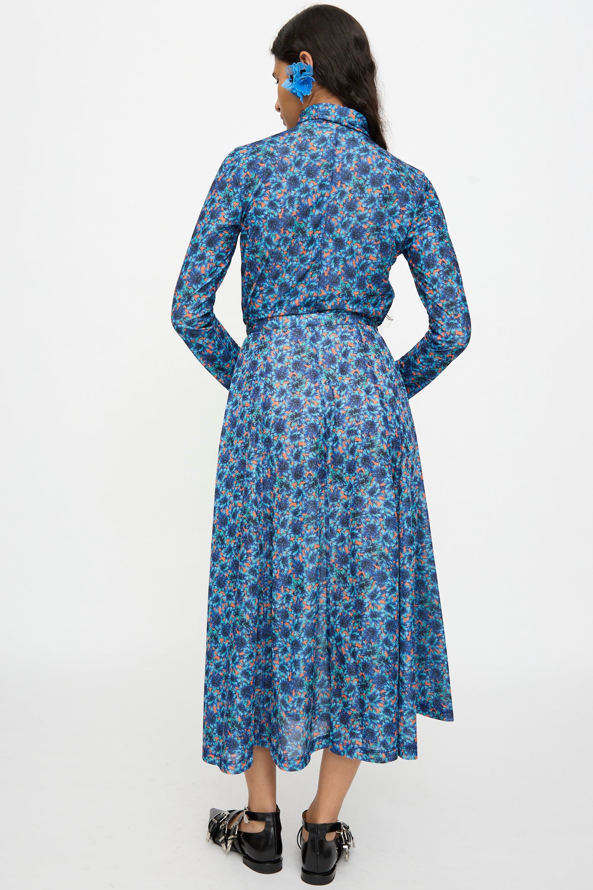 A woman stands facing away against a plain white background, wearing the TOGA ARCHIVES Flared Jersey Dress in Mix—a long-sleeved blue floral print dress featuring an adjustable waist—paired with a matching hair accessory and black shoes.