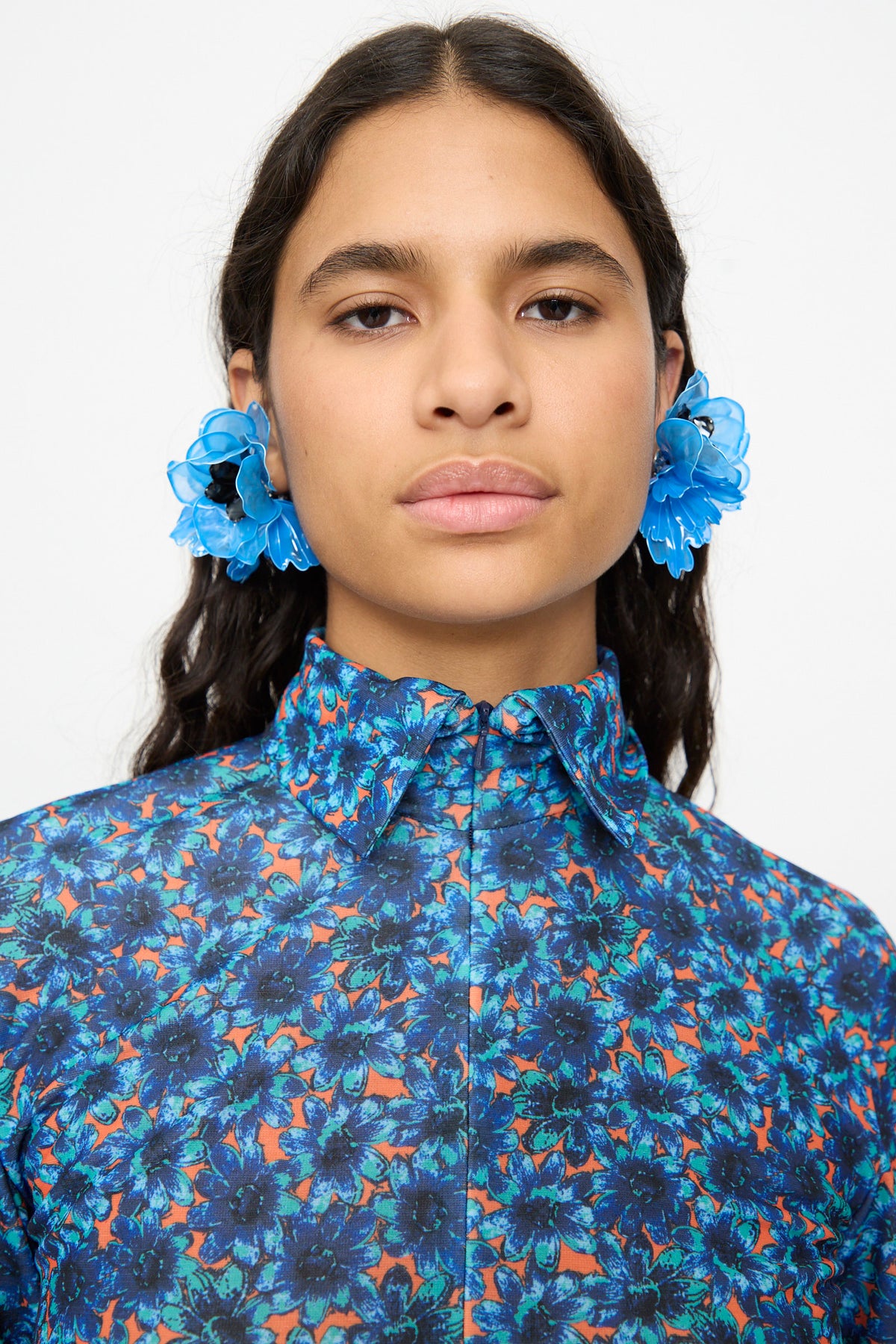 A woman wears TOGA ARCHIVES Flower Earrings in Light Blue—large, flower-shaped clip-on earrings—paired with a blue floral shirt, facing forward with a neutral expression against a plain background.