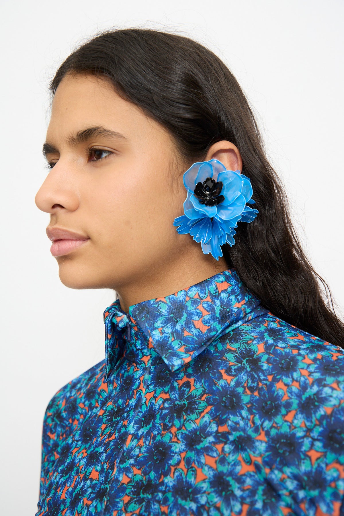 A person with long dark hair is photographed in profile against a plain background, wearing a blue floral-patterned shirt and TOGA ARCHIVES’ Flower Earrings in Light Blue.