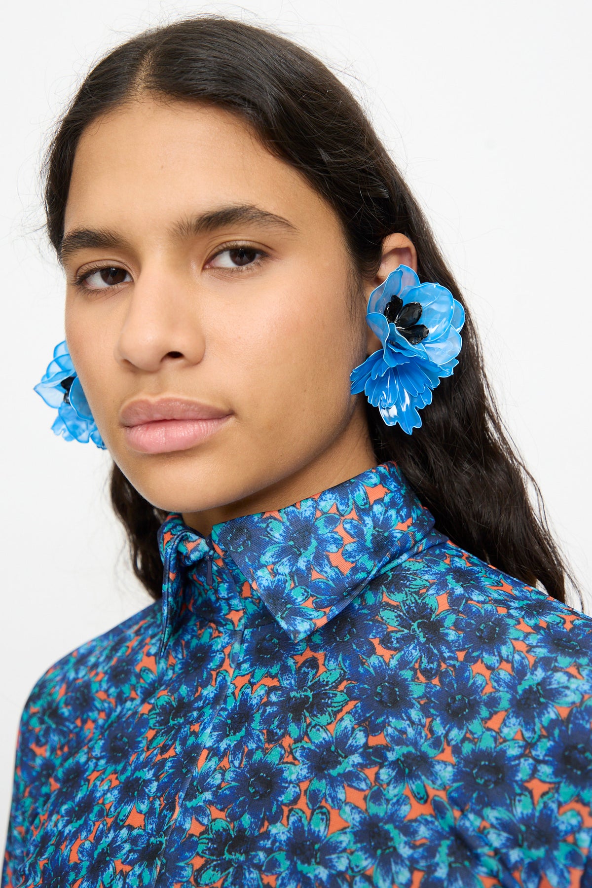 A person with long dark hair wears a blue floral shirt and the TOGA ARCHIVES Flower Earrings in Light Blue, looking at the camera against a plain background.
