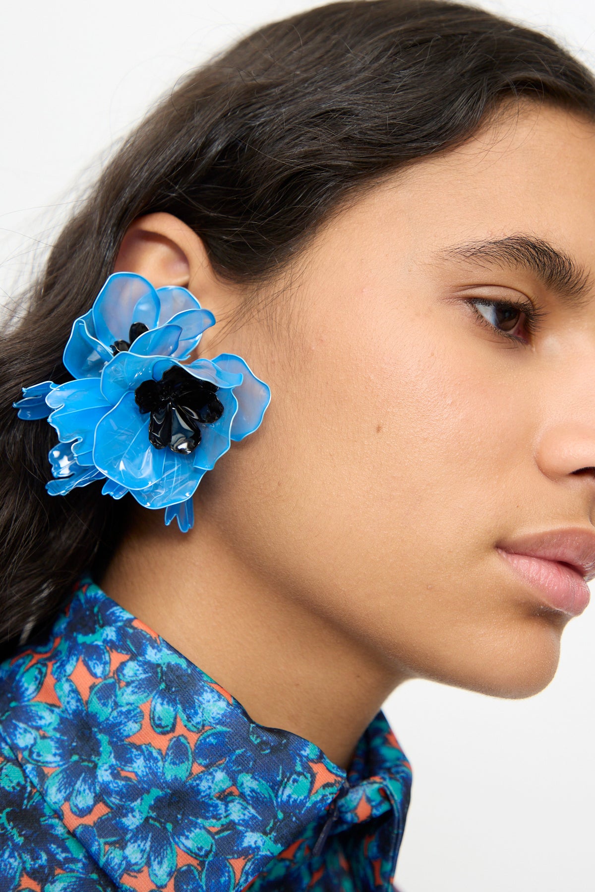 A person wearing the TOGA ARCHIVES Flower Earrings in Light Blue and a blue floral patterned shirt, shown from the side with focus on the earring and part of their face.