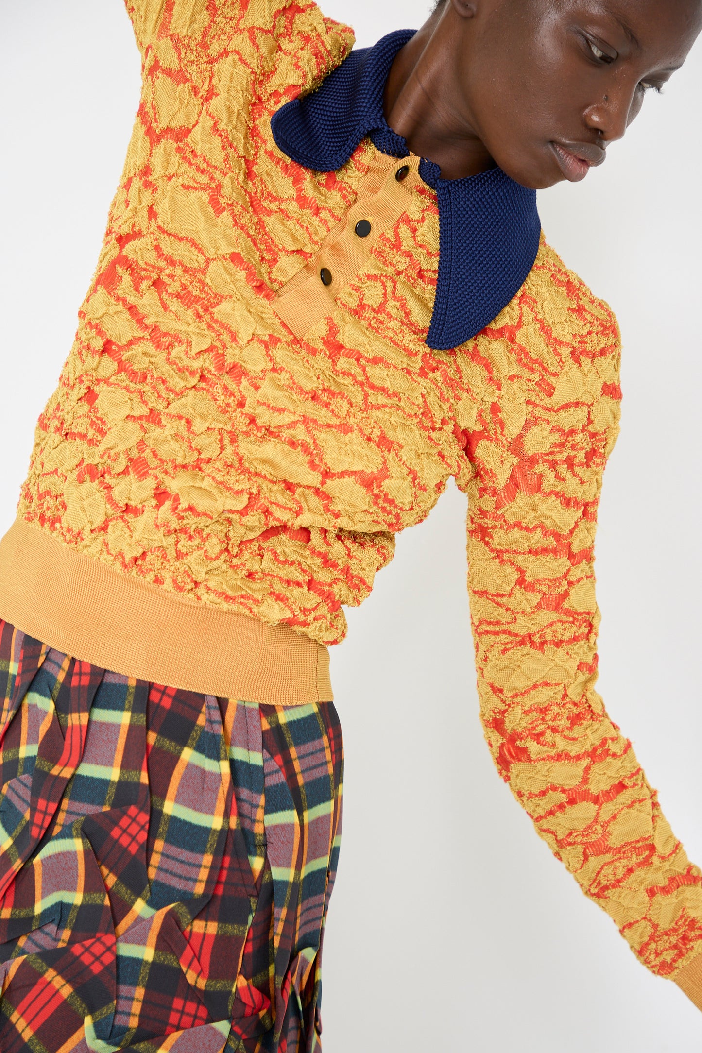 A person is wearing the Flower Jacquard Knit Polo Shirt in Yellow by TOGA ARCHIVES, with a plaid skirt, against a plain background.