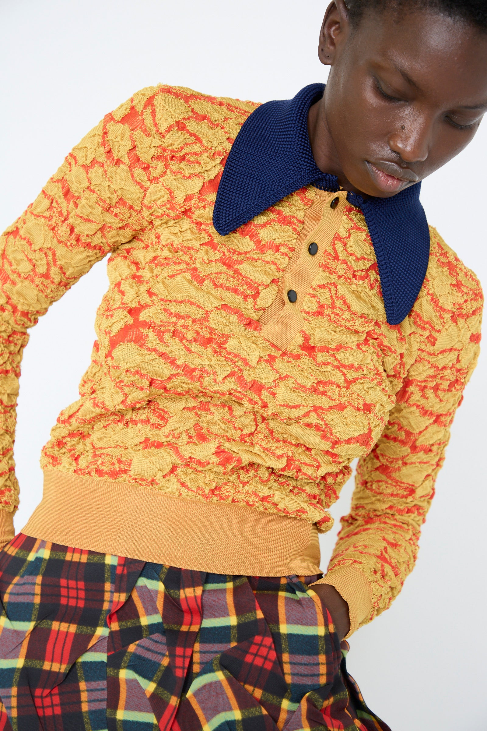 A person wearing the Flower Jacquard Knit Polo Shirt in Yellow by TOGA ARCHIVES, featuring a textured design with a navy collar and long sleeves, paired with plaid skirt, against a plain background.