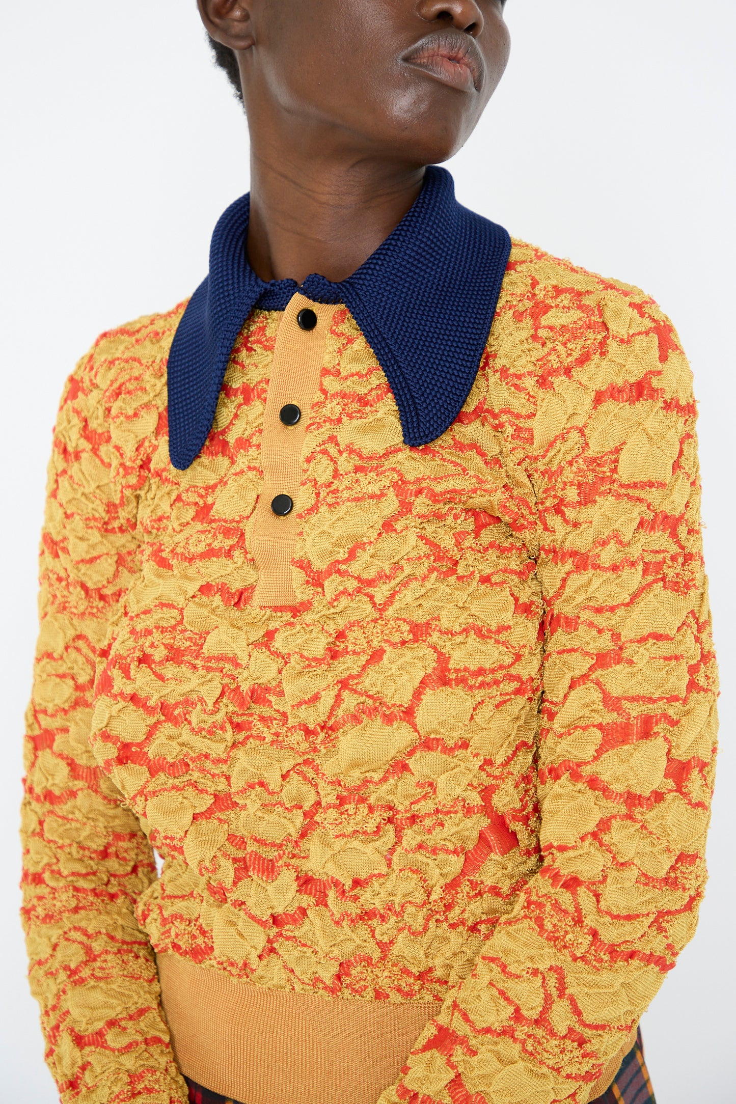 A person is wearing the Flower Jacquard Knit Polo Shirt in Yellow by TOGA ARCHIVES, featuring a textured yellow and orange floral design with a contrasting blue collar and a tan buttoned placket.