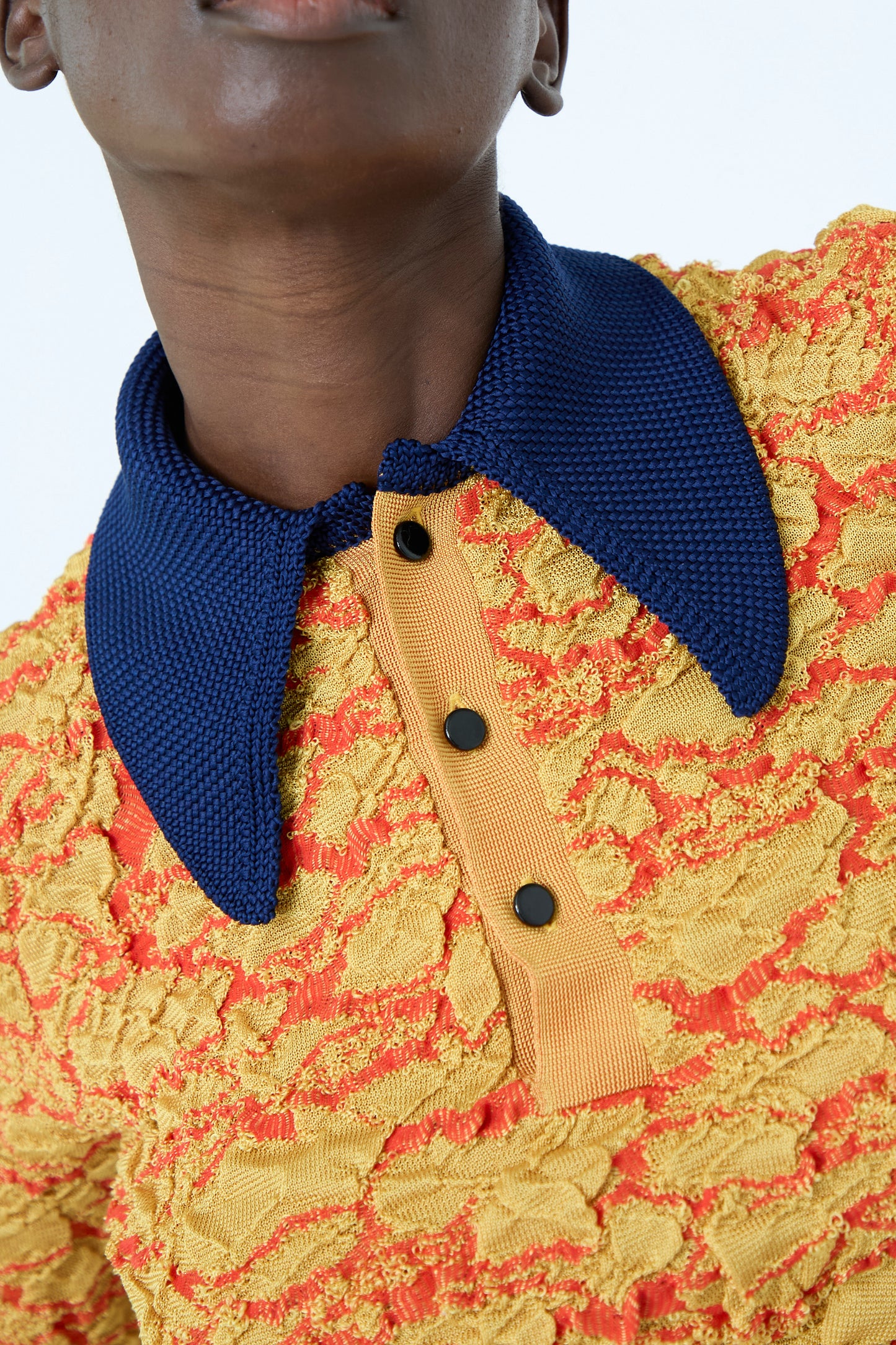 Close-up of an individual wearing the TOGA ARCHIVES Flower Jacquard Knit Polo Shirt in Yellow, featuring a textured floral pattern, a navy blue collar, and buttoned placket.