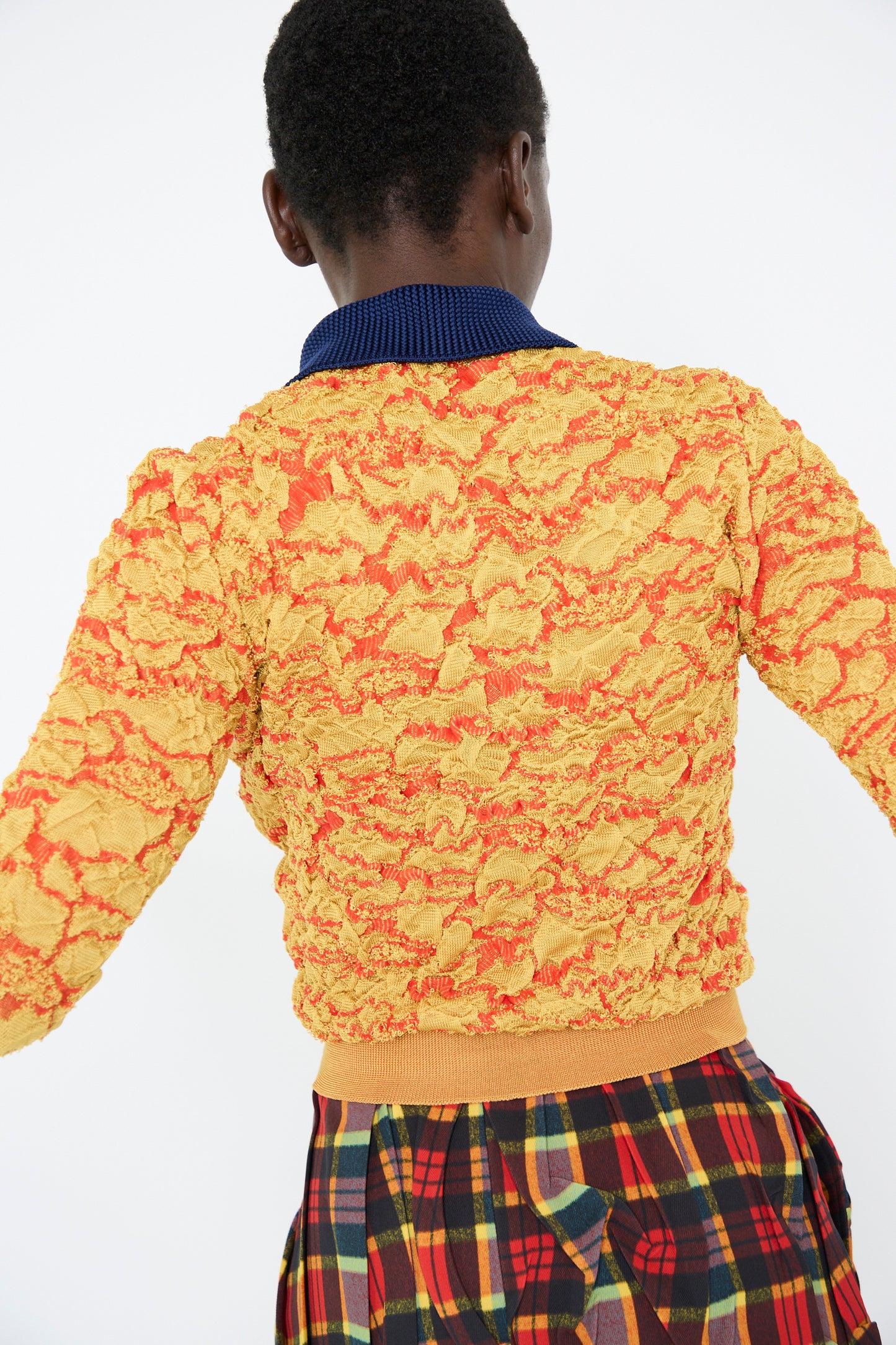 A person wearing the Flower Jacquard Knit Polo Shirt in Yellow by TOGA ARCHIVES, featuring a vibrant yellow and orange texture with a dark blue collar, is paired with a plaid skirt. They face away from the camera in front of a plain white backdrop.