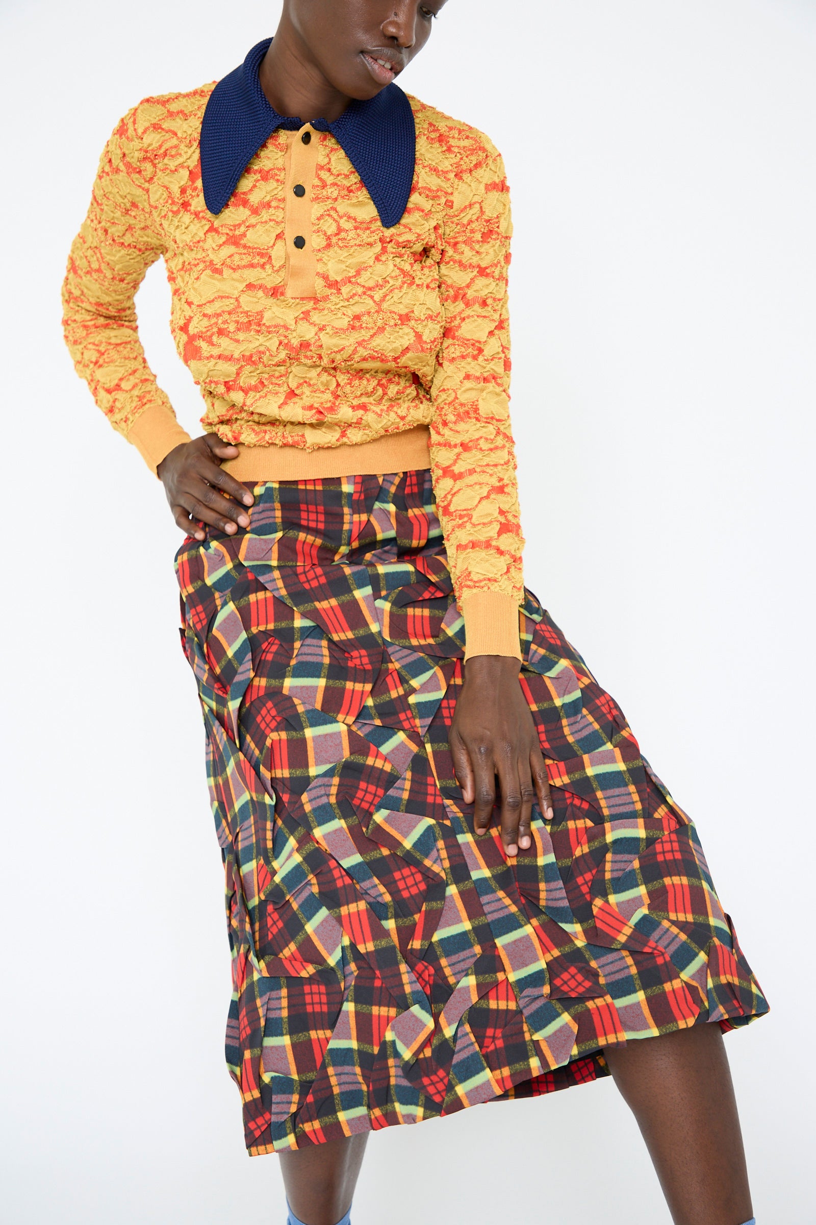 A person is wearing a Flower Jacquard Knit Polo Shirt in Yellow from TOGA ARCHIVES, styled with a colorful plaid skirt, standing against a white background.