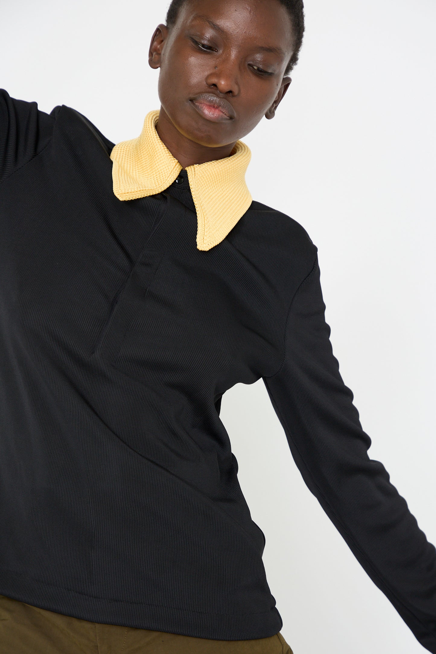 A person wearing a Glossy Rib Polo Shirt in Black by TOGA ARCHIVES, featuring a large yellow knit collar and long sleeves, stands against a white background.