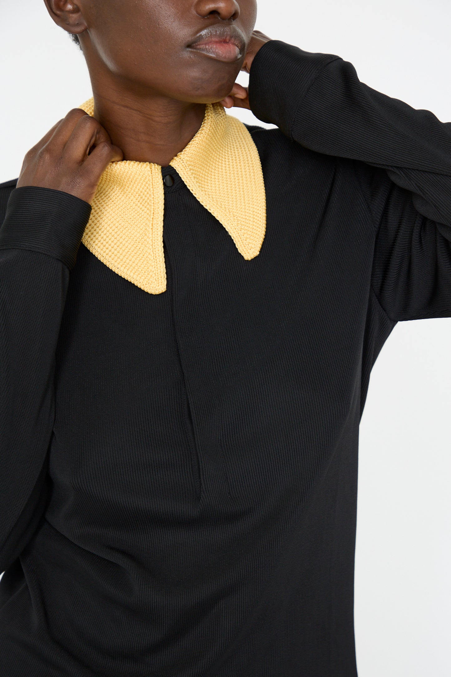 A person wearing the Glossy Rib Polo Shirt in Black by TOGA ARCHIVES, featuring a contrasting yellow knit collar, poses with hands near the collar.