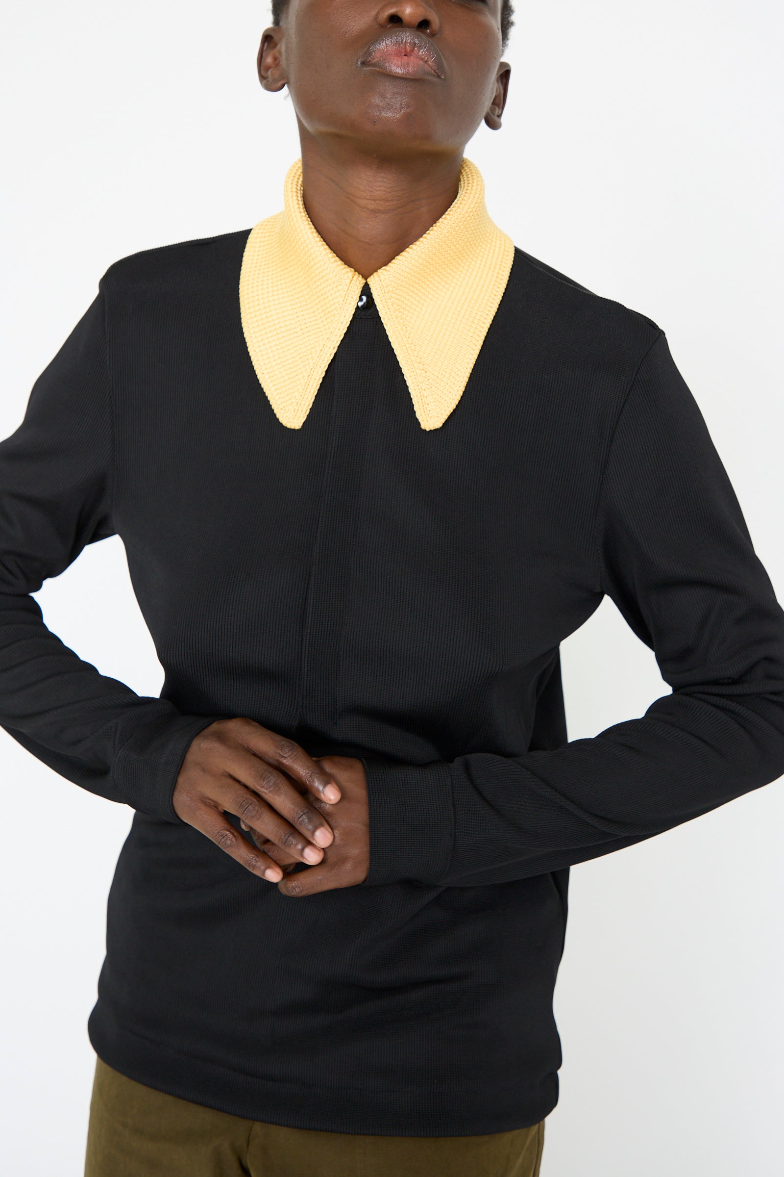 A person wearing the TOGA ARCHIVES Glossy Rib Polo Shirt in Black, featuring a prominent yellow knit collar, stands with hands clasped at the waist against a plain white background.