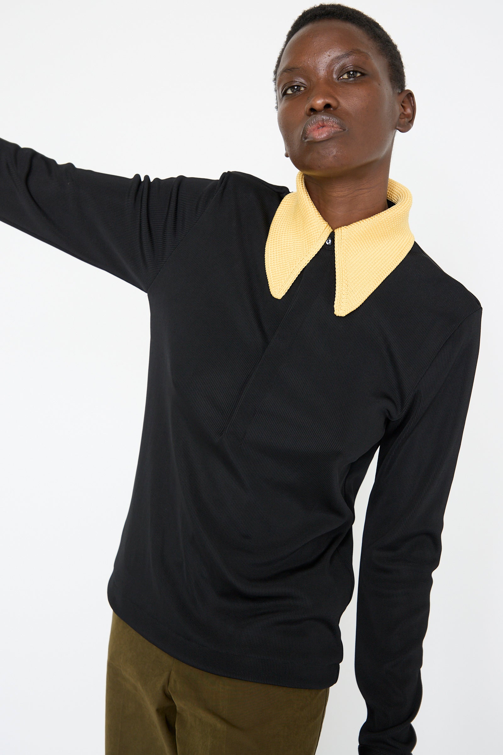 A person wearing the TOGA ARCHIVES Glossy Rib Polo Shirt in Black, which features a distinctive yellow knit collar, stands against a plain background.