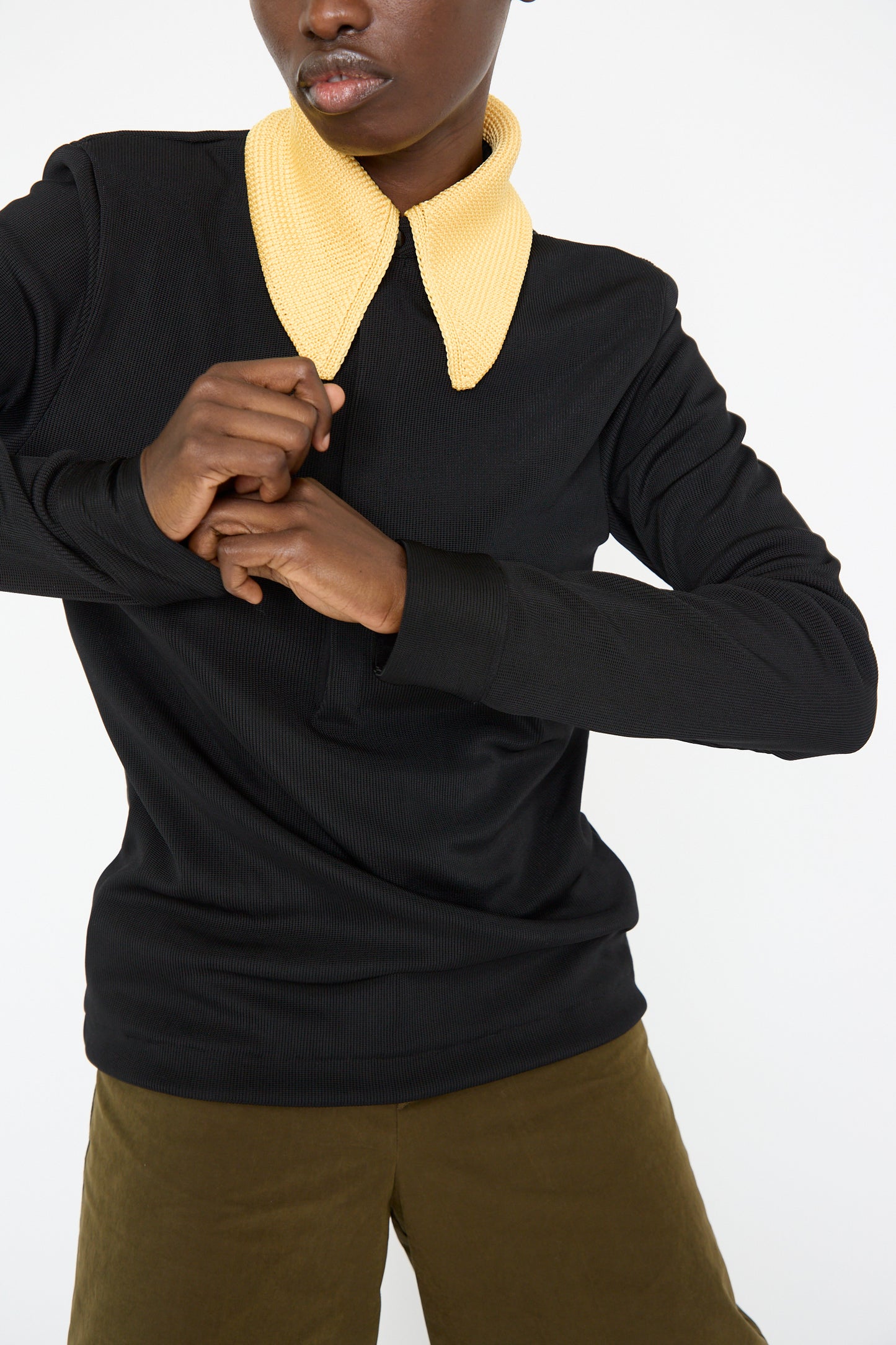 A person wearing the Glossy Rib Polo Shirt in Black by TOGA ARCHIVES, featuring a large, textured yellow knit collar and olive pants, stands against a white background.