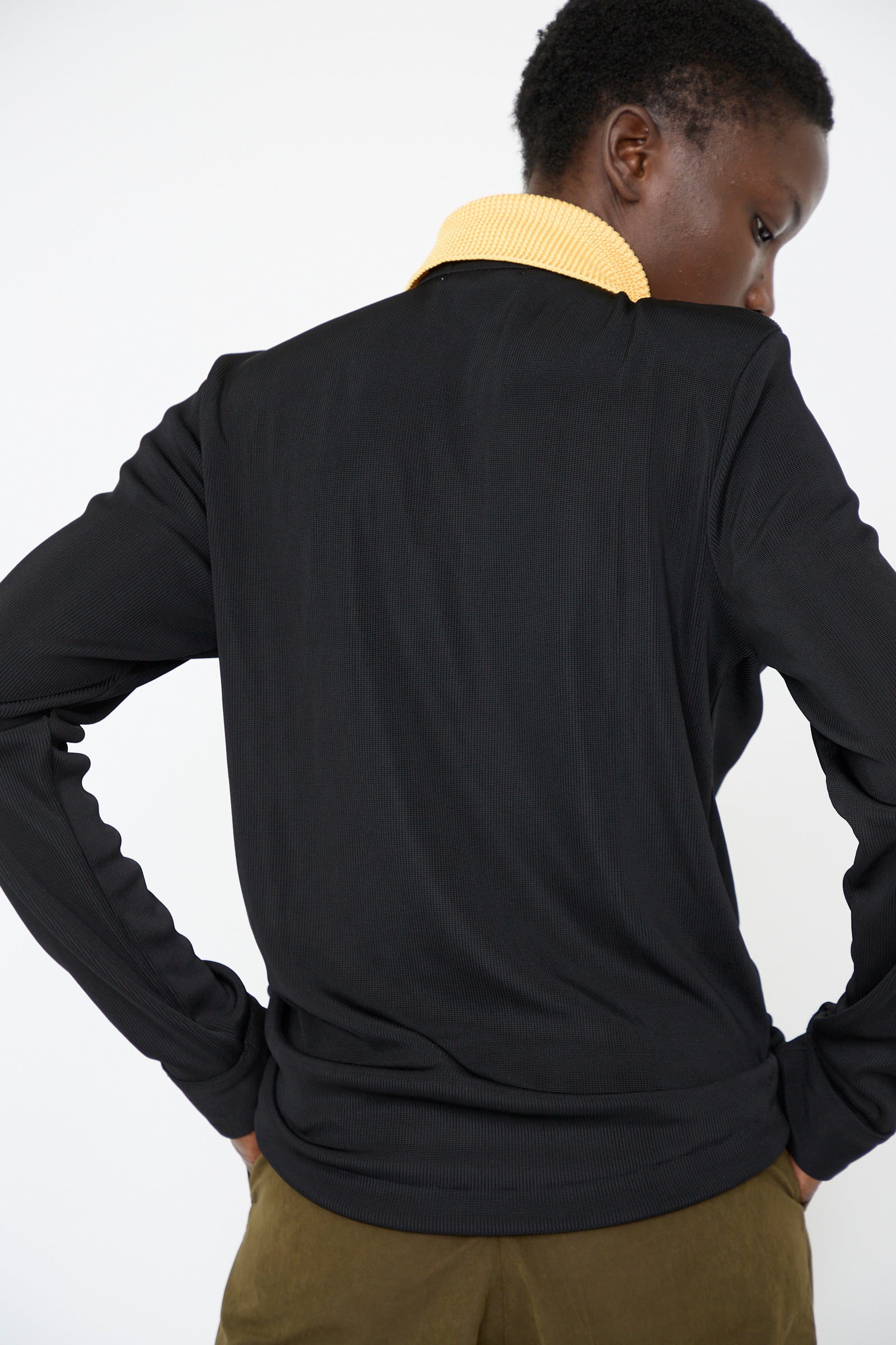 A person wearing the Glossy Rib Polo Shirt in Black by TOGA ARCHIVES, featuring a yellow knit collar and long sleeves, stands with their back to the camera, hands on hips.