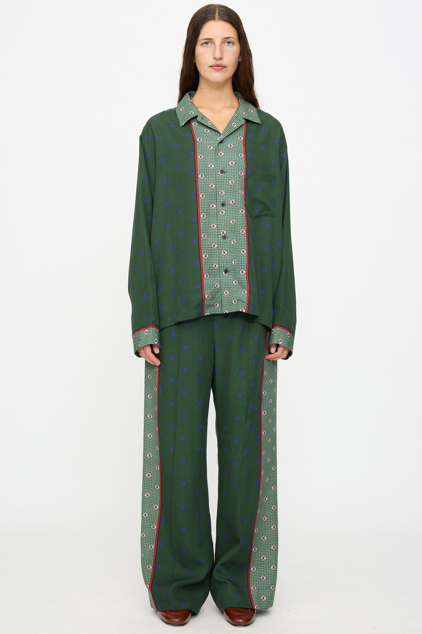 A person stands against a white background wearing the TOGA ARCHIVES Inner Print Drawstring Pant in Green, featuring a loose, green floral pattern with red and blue accents, styled with a matching pajama-style shirt and brown shoes.