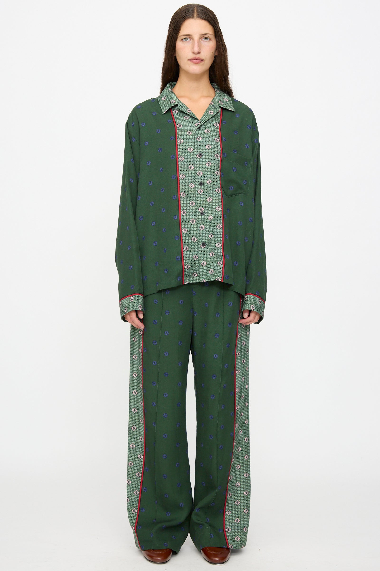 A person stands against a white background wearing the TOGA ARCHIVES Inner Print Drawstring Pant in Green, featuring a loose, green floral pattern with red and blue accents, styled with a matching pajama-style shirt and brown shoes.