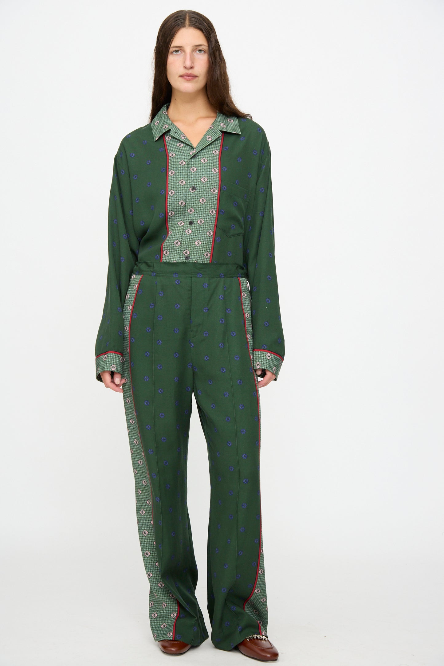A person stands against a plain background wearing the TOGA ARCHIVES Inner Print Drawstring Pant in Green, featuring a matching green patterned shirt with red accents and brown shoes.