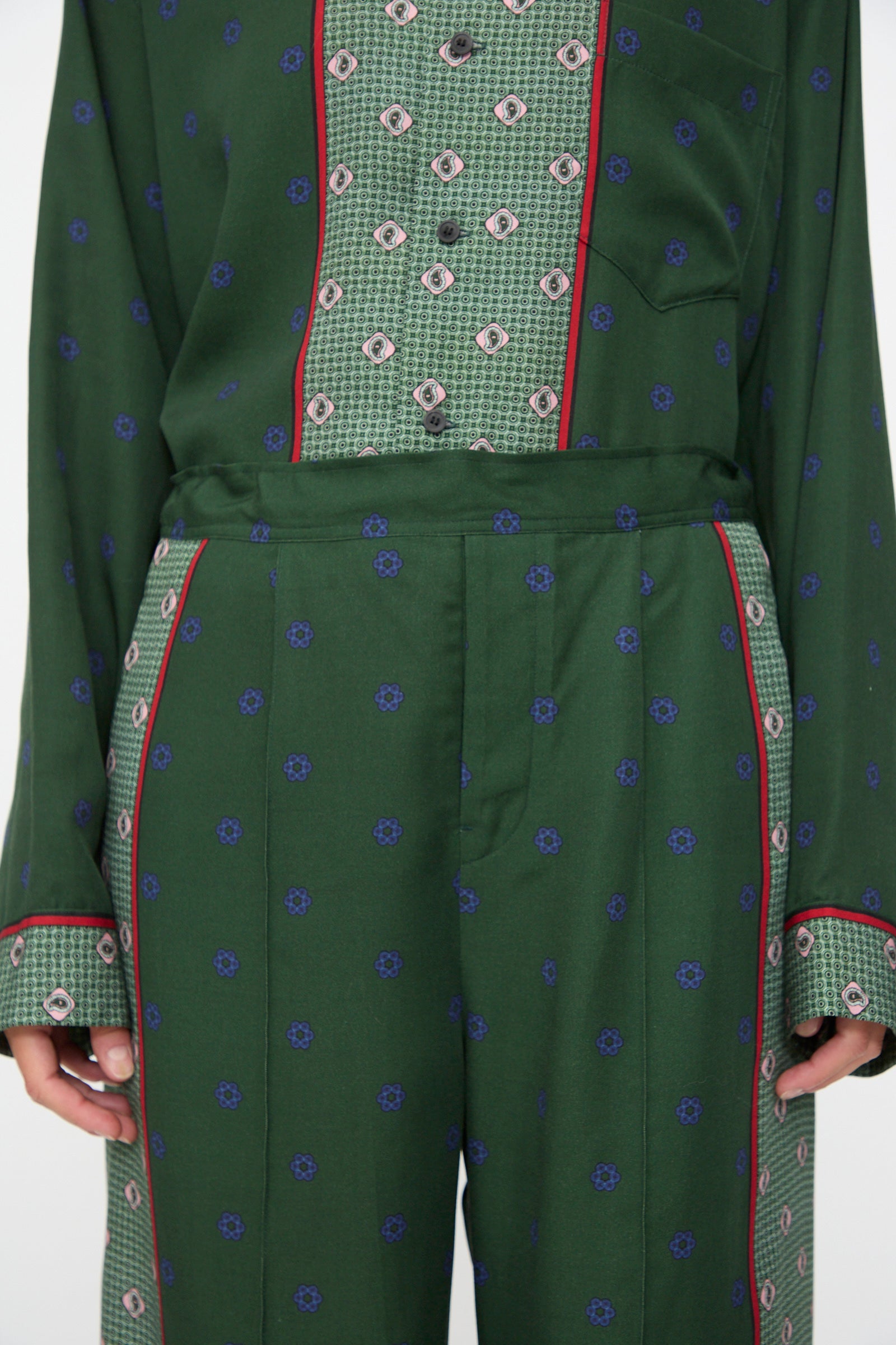 A person is wearing the TOGA ARCHIVES Inner Print Drawstring Pant in Green, featuring geometric designs, blue floral motifs, and red piping, paired with a matching green patterned shirt.